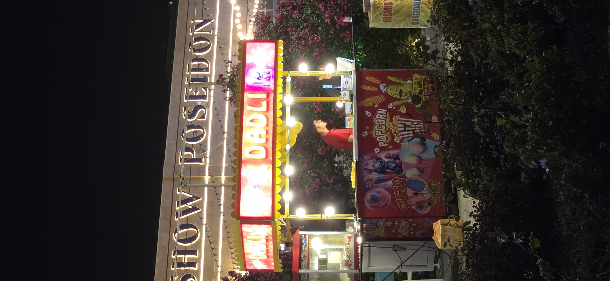 The image shows a night scene at a street food vendor in Sumgayit, Azerbaijan. A brightly lit sign above reads "SHOW POSEIDON." Below this, a food stall is set up, featuring a prominent neon sign that says "DADLI" in both Turkish and Azerbaijani languages, meaning "delicious." The stall appears to sell popcorn and cotton candy, with graphics on its facade depicting popcorn bags and cotton candy. A person in a red shirt is visible behind the counter, possibly serving customers, and another person is partially visible to their right. To the left of the stall, a popcorn machine is present. The surrounding area is dark, with illuminated lights on the stall and foliage with pink flowers visible in the background, suggesting it's an outdoor setting. The overall impression is of a lively night market or fairground.