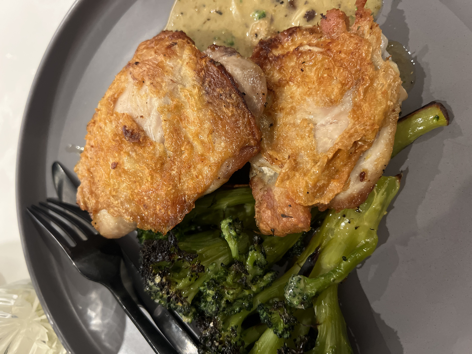 The image shows a close-up of a plate of food. Two pieces of golden-brown, crispy-skinned chicken are perched on top of a bed of roasted broccoli. A creamy, pale-yellow sauce is drizzled near the chicken. A dark grey fork rests on the left side of the plate. The plate itself is a matte grey. The background shows the edge of a white table and what appears to be a white flower.