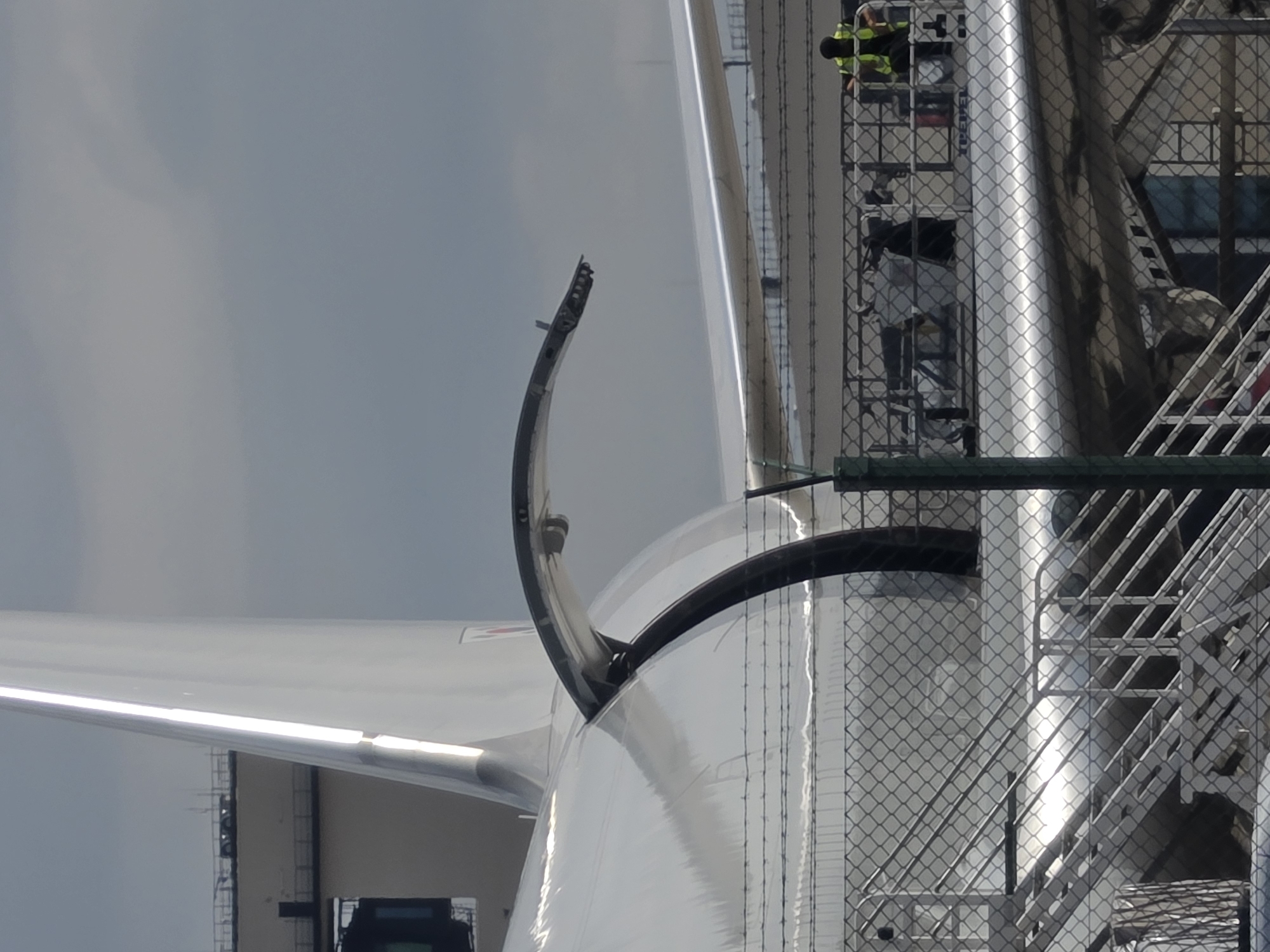 A close-up, slightly tilted, eye-level shot captures the tail section and rear fuselage of a large, white aircraft parked on an airport tarmac under a cloudy sky. A dark, curved panel is partially open, revealing mechanical components. A chain-link fence with barbed wire on top runs across the foreground, obscuring a portion of the aircraft's lower fuselage and stairs. In the middle ground, behind the fence, a maintenance platform is positioned alongside the aircraft. On the platform, two individuals are visible: one wearing a high-visibility yellow vest and dark pants, and another person in dark clothing. The environment appears to be an operational area of an airport, likely Frankfurt Airport, Germany, given the presence of aircraft and maintenance equipment.