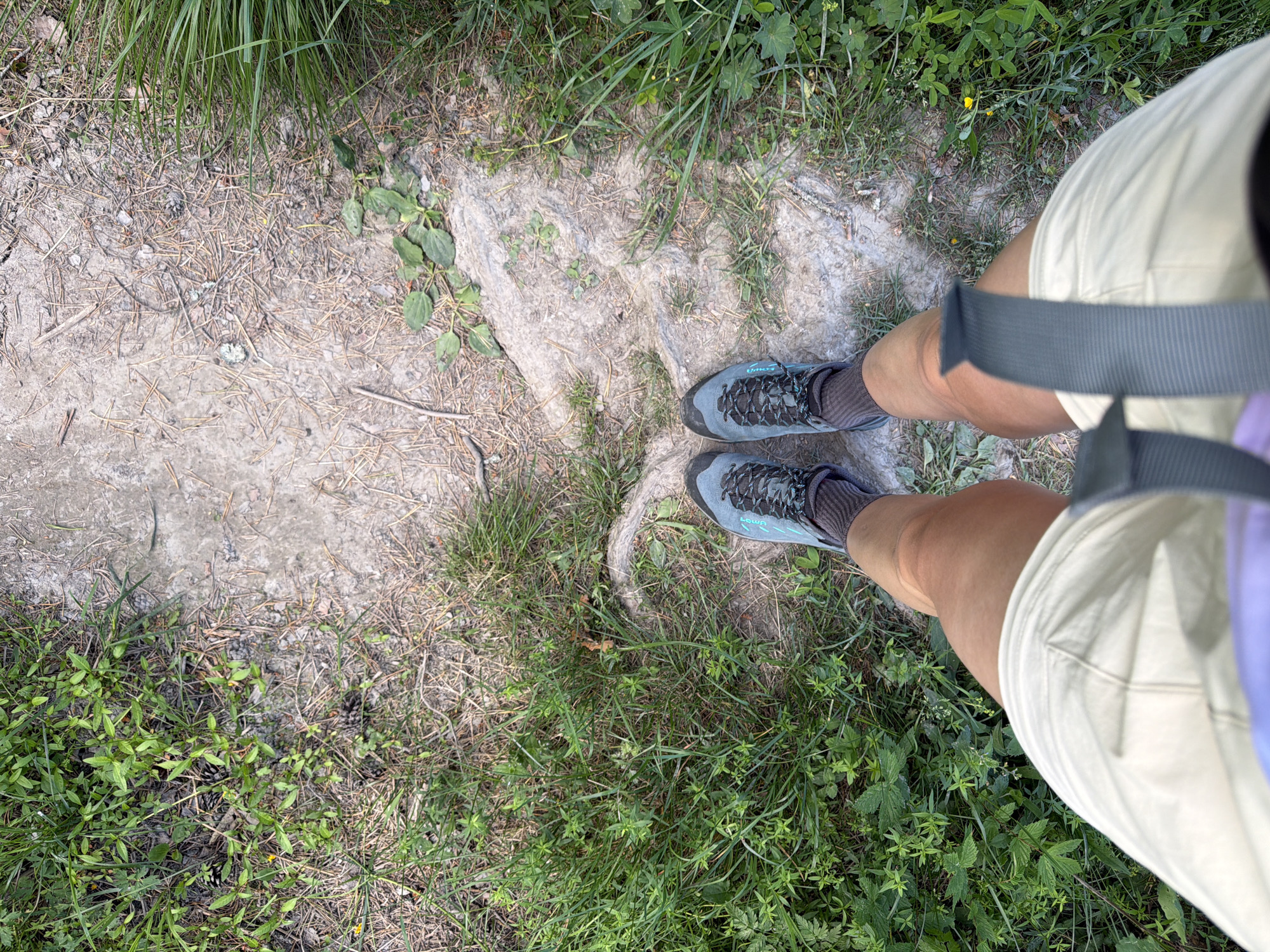 An overhead shot shows a pair of legs wearing light-colored shorts and hiking boots standing on a dirt path surrounded by grass and pine needles. The path appears to be in a natural setting, possibly a park or forest. The person is standing still, and the image focuses on their lower body and the immediate surroundings. No specific landmarks or identifying features of Kuznechnoye, Russia, are visible.