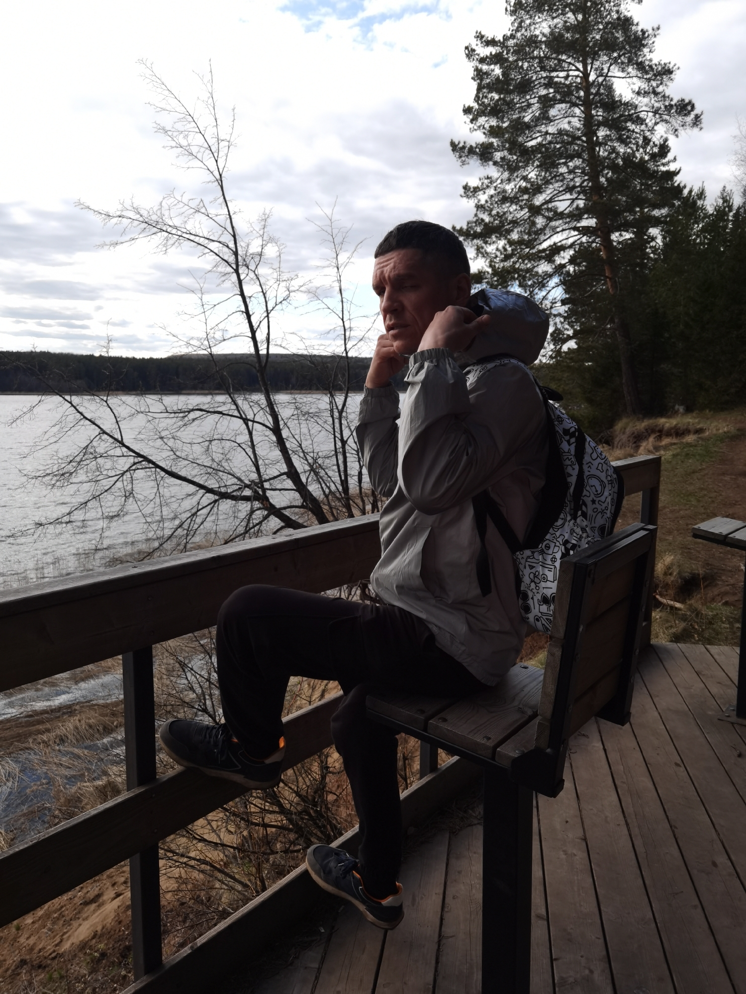 A man is sitting on a wooden bench at an outdoor overlook. The man has dark hair and is wearing a grey jacket, black pants, and black shoes with orange accents. He is pulling up the hood of his jacket and has a backpack on his back. He is looking to the left of the frame. In the background, there is a lake, trees, and a cloudy sky. The overlook has a wooden railing and deck. A second bench and table are to the right. The location is most likely Votkinsk, Russia, a city known for its reservoir.