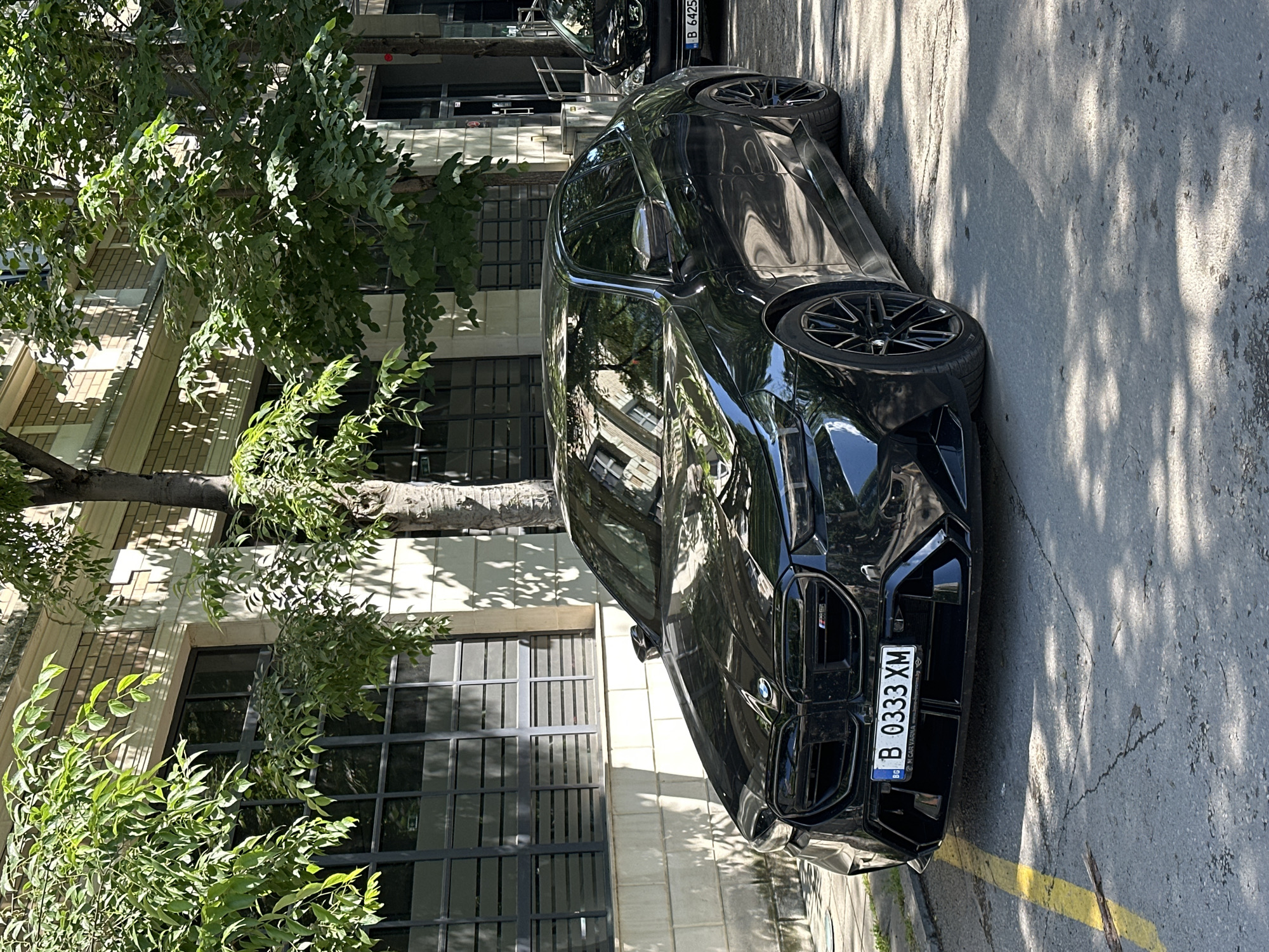 A sleek, black BMW XM is parked on a street in Varna, Bulgaria. The car's front is angled towards the viewer, displaying its distinctive kidney grille and license plate "B 0333 XM." The vehicle is positioned adjacent to a yellow painted line marking the parking space. Behind the car, a building with large, multi-pane windows is visible, partially obscured by lush green foliage from a tree lining the sidewalk. Another vehicle is parked in the background. The scene is bathed in natural daylight.