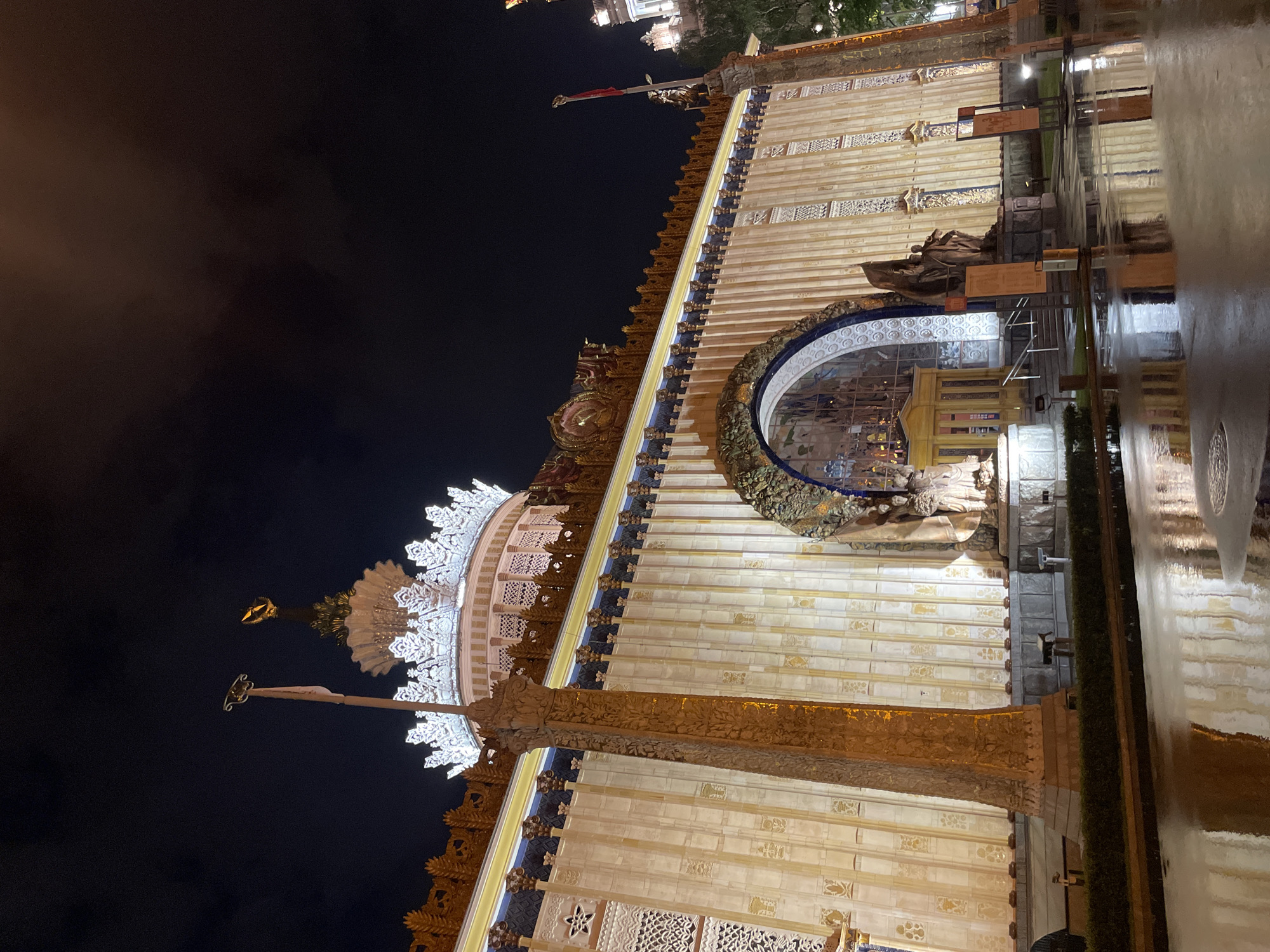 The image depicts the facade of the Pavilion No. 59 "Grain" at the VDNKh (Exhibition of Achievements of National Economy) in Moscow, Russia, at night. The pavilion is elaborately decorated with ornate architectural details, including sculptures and intricate patterns. The building is brightly illuminated, contrasting with the dark night sky. The foreground shows a wet surface reflecting the building's lights, suggesting it has recently rained. There are no people visible in the frame. A flag pole is on top of the pavilion.