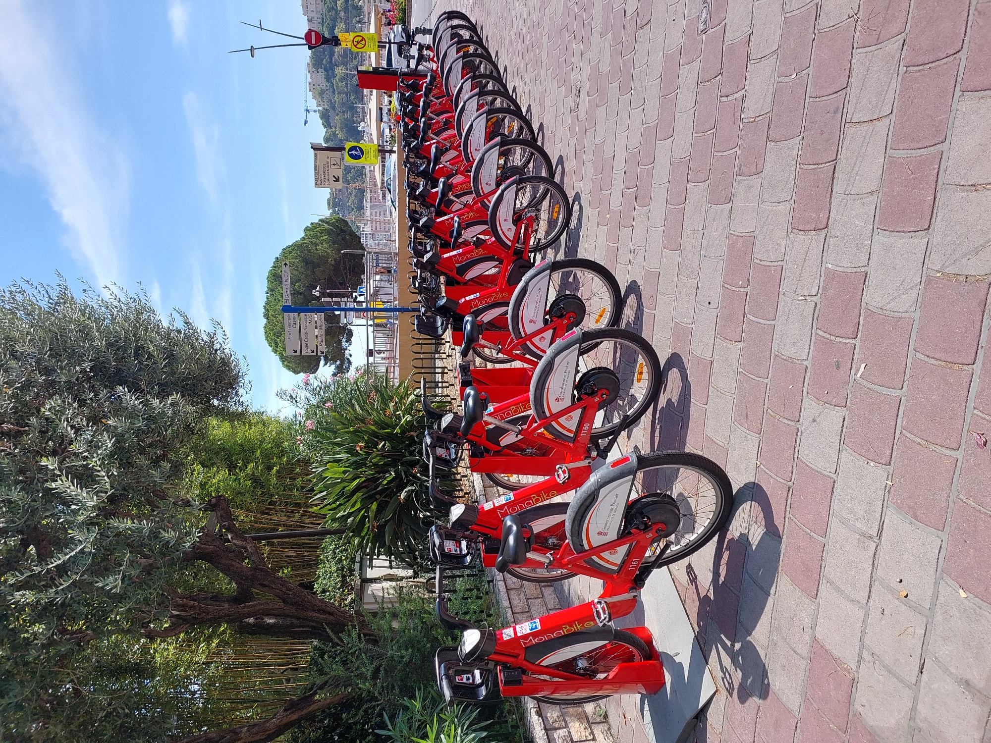 A line of red MonaBike rental bicycles is parked on a brick-patterned sidewalk in Monaco. The bikes are evenly spaced and have digital displays on the handlebars. In the background, there is lush green foliage, including bamboo and palm trees, and a blue sky with scattered clouds. Signage in the background indicates directions to Port Hercule, Casino, Gare S.N.C.F., and Palais-Musées, suggesting a location near the port and city center. A traffic light and pedestrian crossing signs are also visible. The scene captures a sunny day in a public area of Monaco.