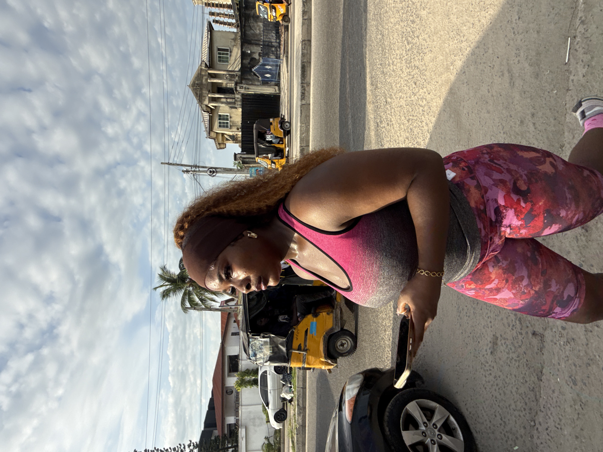 A woman stands on a paved surface next to a car's front tire. She is wearing a pink and gray sports bra and floral leggings. She is holding a cell phone. Behind the car is a yellow auto rickshaw. In the background are buildings and a cloudy sky. The location appears to be a street in Ado, Nigeria.