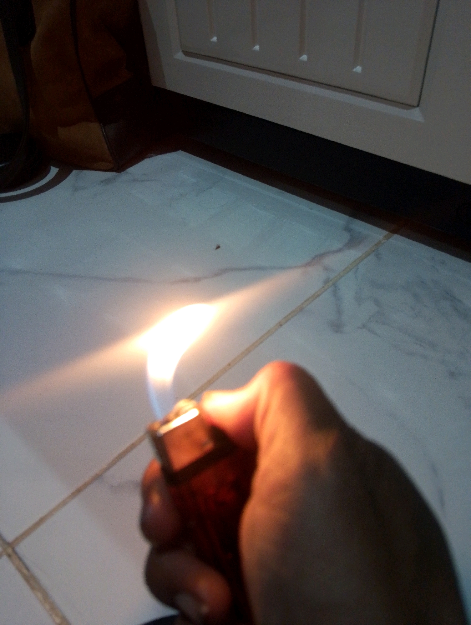 In Sumbergirang, Indonesia, an individual's hand is shown holding a red lighter with a flame emanating from its tip. The lighter is positioned in the foreground, with the flame casting a bright orange and white glow. The background features a tiled floor with a marbled pattern and a white cabinet or appliance. The lighting is dim, with the primary light source appearing to be the flame of the lighter.