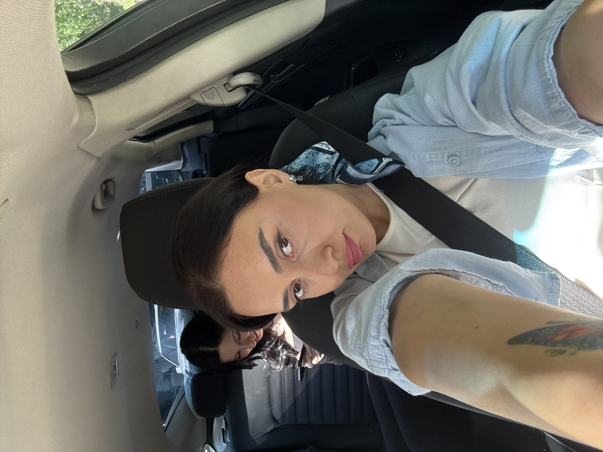 A selfie-style portrait, taken inside a car during daytime in Tbilisi, Georgia, features two women. The woman in the foreground, with dark hair, dark eyebrows, and pink lipstick, looks directly at the camera with a neutral expression. She wears a light-colored top under a light blue collared shirt, with a black seatbelt crossing her chest. A blue and white patterned scarf or bandana is visible near her shoulder, featuring partially legible text that starts with "Blueb...". She also wears a small, ornate earring and has a colorful butterfly tattoo on her upper arm.
In the background, another woman with dark hair is seen resting with her head against a dark headrest, appearing to be asleep or relaxed. She holds a dark-colored smartphone in her hand.
The car's interior is predominantly light-colored, with a beige headliner and A-pillar. An open sunroof is visible, allowing bright natural light into the vehicle, suggesting a sunny day. A seatbelt mechanism includes a partially visible "AIRBAG" label. The car seats are dark. The image is composed from an angled, slightly upward perspective, characteristic of a handheld selfie.
