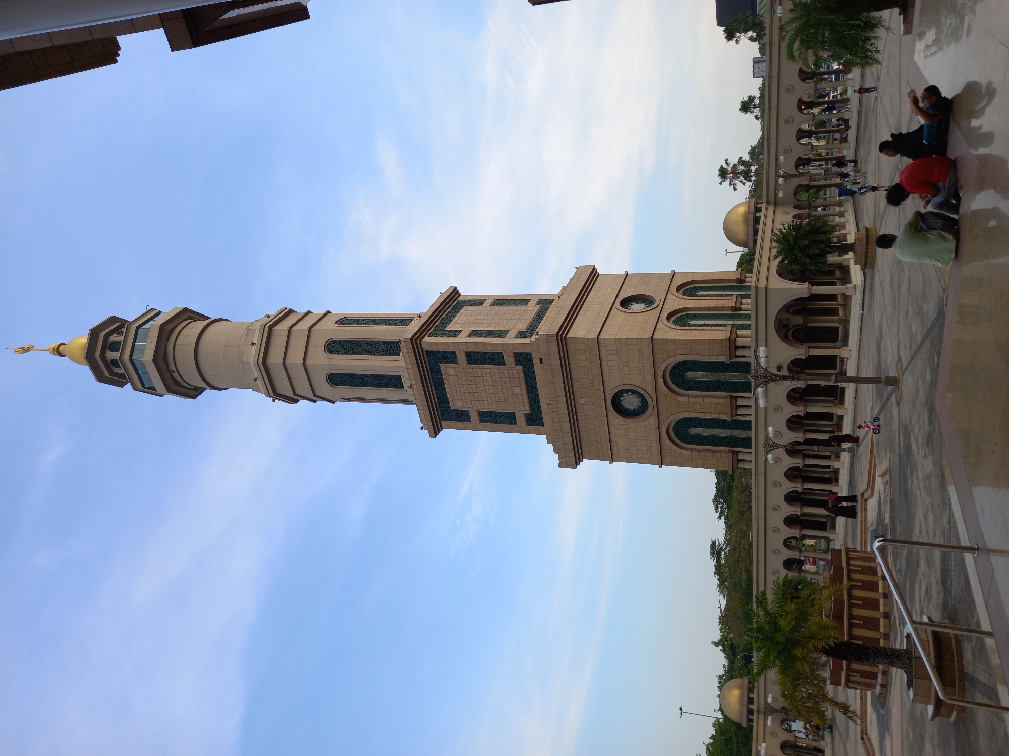 The image depicts the majestic Baitul Muttaqien Grand Mosque in Samarinda, Indonesia, with its towering minaret dominating the skyline. The minaret, a beige structure with green accents, is topped with a golden dome. In the foreground, a paved courtyard is visible, with several people seated and lying down on the reflective floor. Some individuals are casually dressed, while others wear traditional Islamic attire. The mosque's facade, featuring arched walkways and decorative elements, is seen in the background. The sky is a soft blue with wispy clouds. The overall scene conveys a sense of peace and tranquility within the mosque's premises.