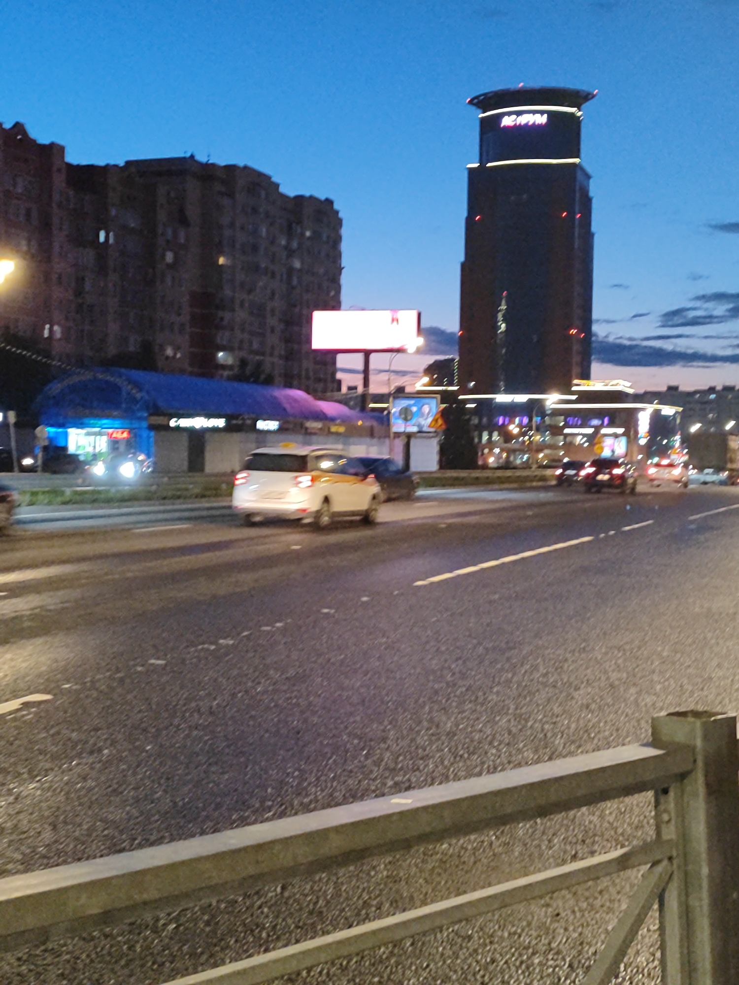 The image depicts a street scene at dusk in an urban environment. A multi-lane road takes up the majority of the foreground and midground, with visible lane markings. Several cars are present on the road, including a white taxi with yellow markings. In the background, a tall, dark skyscraper with a distinctive cap is illuminated by lights and signage, one of which appears to read "ACRYM". To the left of the skyscraper, a large, multi-story residential building is visible. Further left, a blue structure, possibly a car wash or service area, is brightly lit with blue and white lights. The sky is a deep blue, transitioning from twilight, with some clouds present. A metal guardrail runs along the bottom right of the image. The overall impression is of a busy city street at the end of the day.