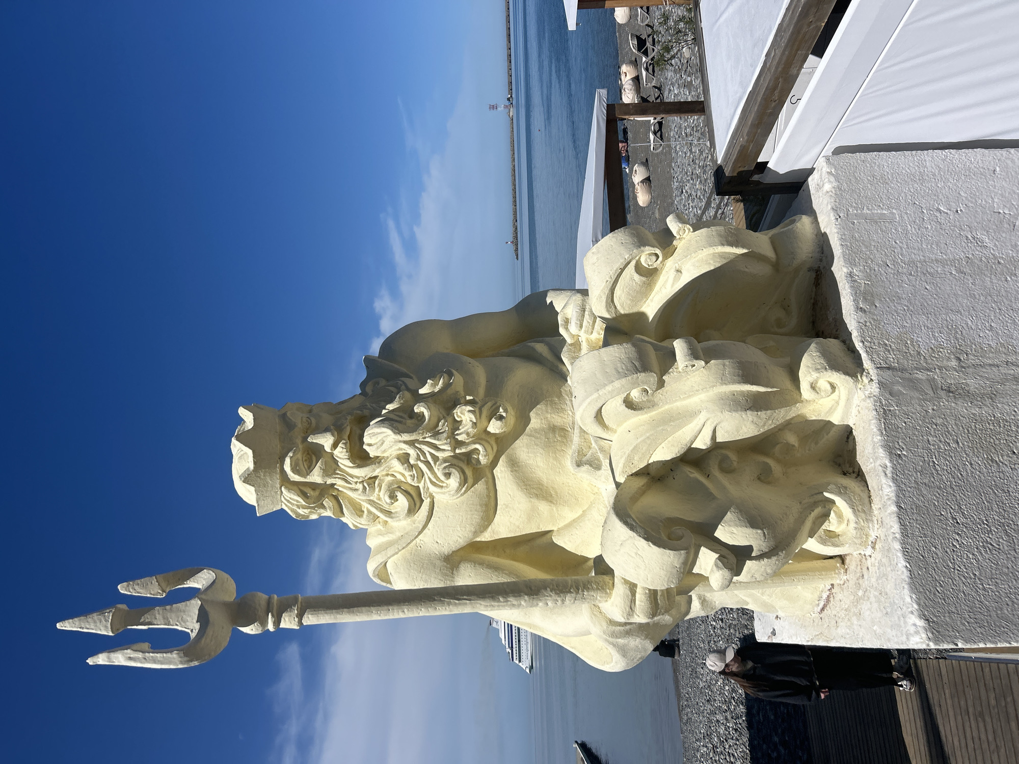 The image depicts a light-colored, likely concrete, statue of Neptune holding his trident. The statue is situated on a raised platform with a white concrete wall. Behind the statue, there is a pebble beach and the Black Sea. In the distance, a breakwater and possibly a distant structure are visible, as well as a large ship. Above the statue, the sky is blue with scattered clouds. The statue is positioned on a deck, with a white fabric awning nearby, indicating a restaurant or lounge area on the beach. In the background there are beach loungers. A person dressed in black stands at the base of the statue. The location is most likely in the city of Sochi, Russia.
