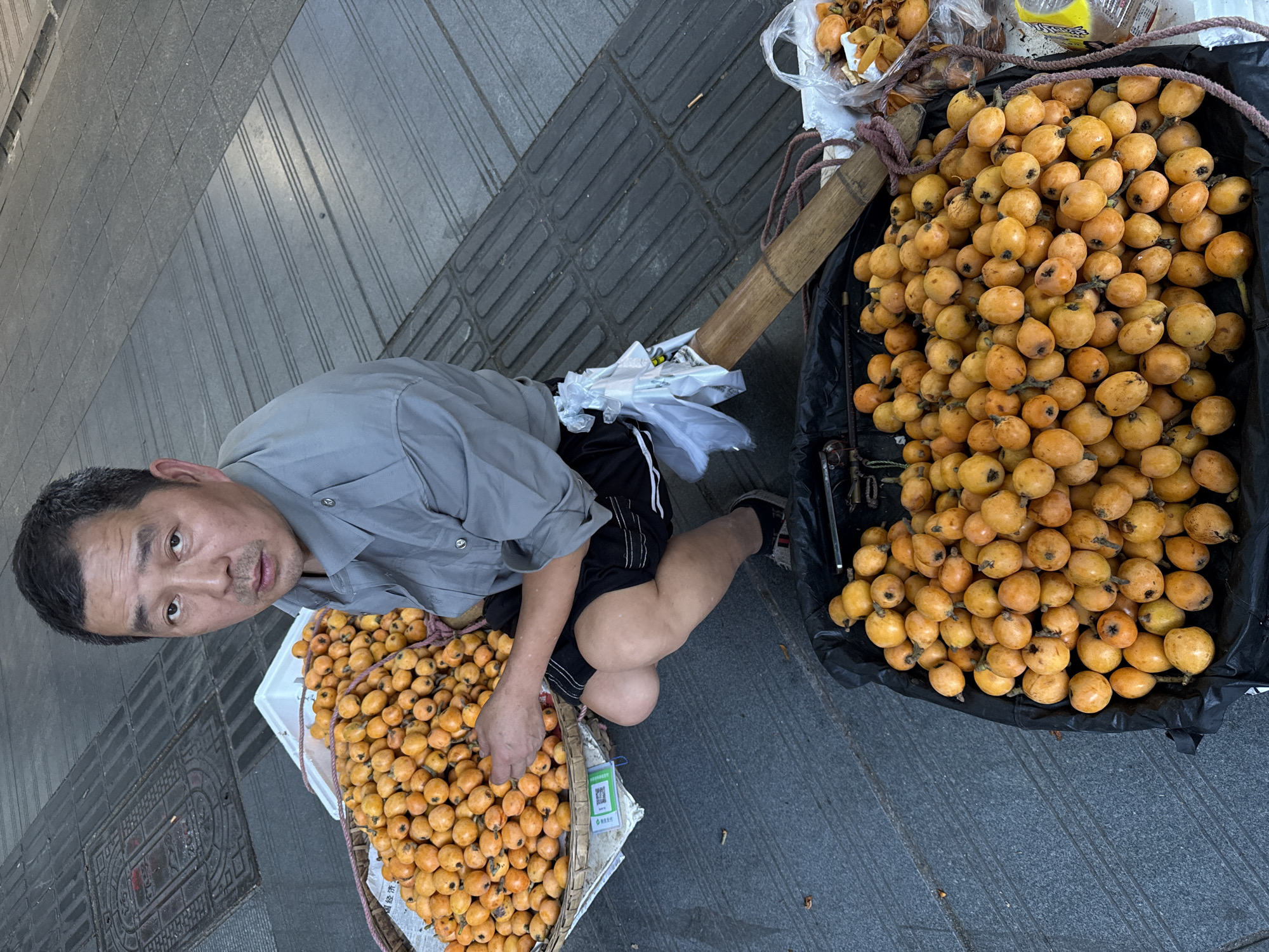 An eye-level, medium shot shows a Chinese man selling loquats (also known as Japanese plums) on the street in the city of Beibei, China. He is kneeling on the gray sidewalk in front of two baskets of fruit. The man is light-skinned, with black hair that is receding at the temples, dark eyes, and a slight mustache. He is wearing a gray short-sleeved shirt and black shorts with white trim. He is looking at the viewer. To the man's left, in a white woven basket, are numerous loquats. His hand is resting on the fruit. Behind him and to the right, another black basket overflows with loquats. A bamboo pole rests across the two baskets, presumably for carrying the goods. In the background, the sidewalk is made of gray paving stones.