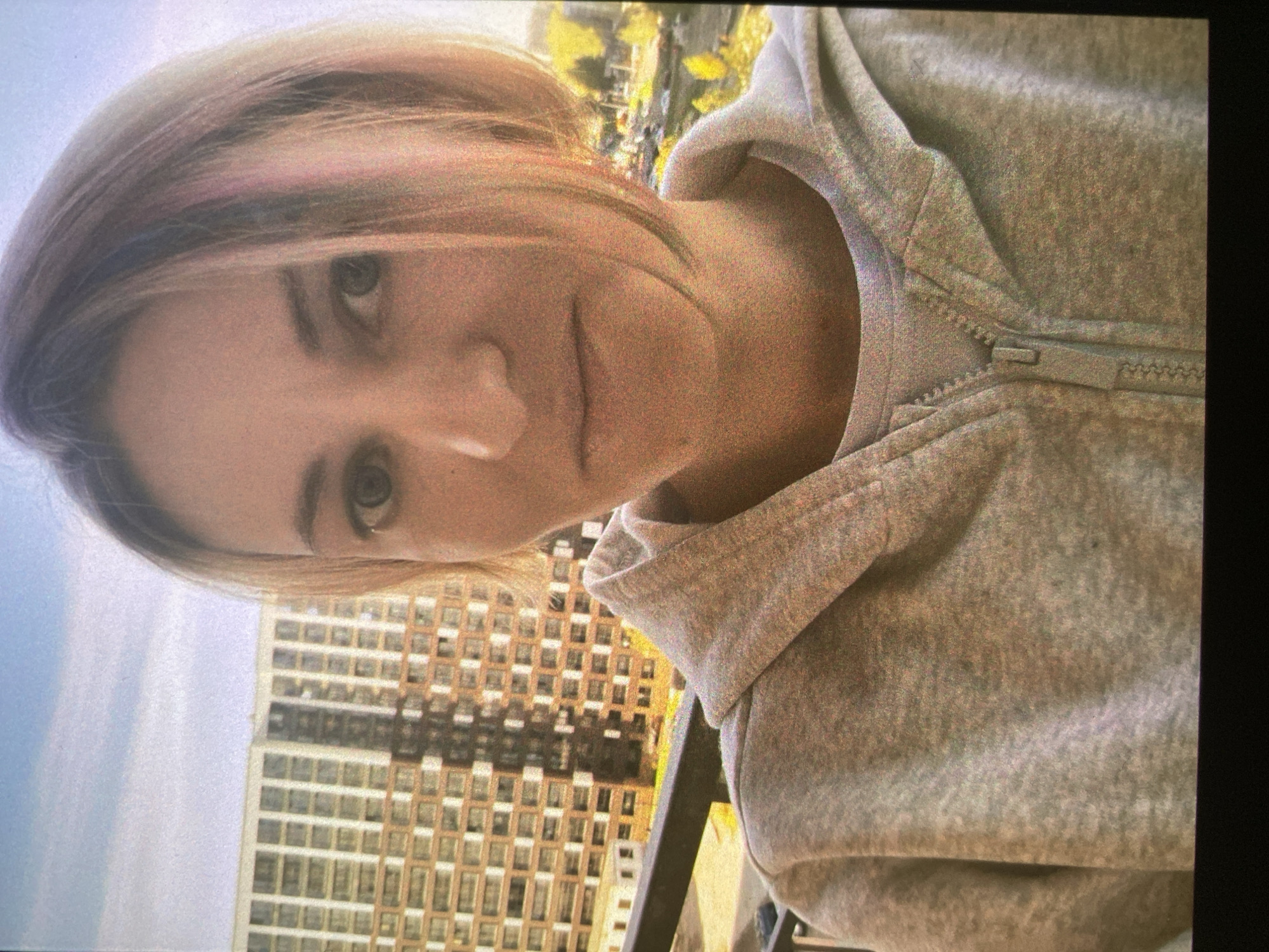 A selfie, rotated 90 degrees clockwise, captures a person, likely female, with light-colored hair that shows subtle pinkish and purplish highlights. She is looking directly at the camera with a neutral to slight smile, her face illuminated by natural light. She wears a light gray hooded sweatshirt with a visible zipper partially open at the neck.
The setting appears to be outdoors, possibly on a balcony or at a high elevation, with a clear or lightly clouded sky in the upper left background (when oriented correctly). Behind the person, a prominent feature is a tall, multi-story building characterized by a repetitive grid pattern of numerous windows. Beyond the building, hints of trees with vibrant yellow and orange foliage suggest an autumn season. The lighting appears to be daytime, possibly late afternoon, casting soft tones on the subject and background. The image has a somewhat grainy texture. No visible text is present. The general urban landscape with residential buildings and autumnal colors is consistent with the provided context of Saint Petersburg, Russia.