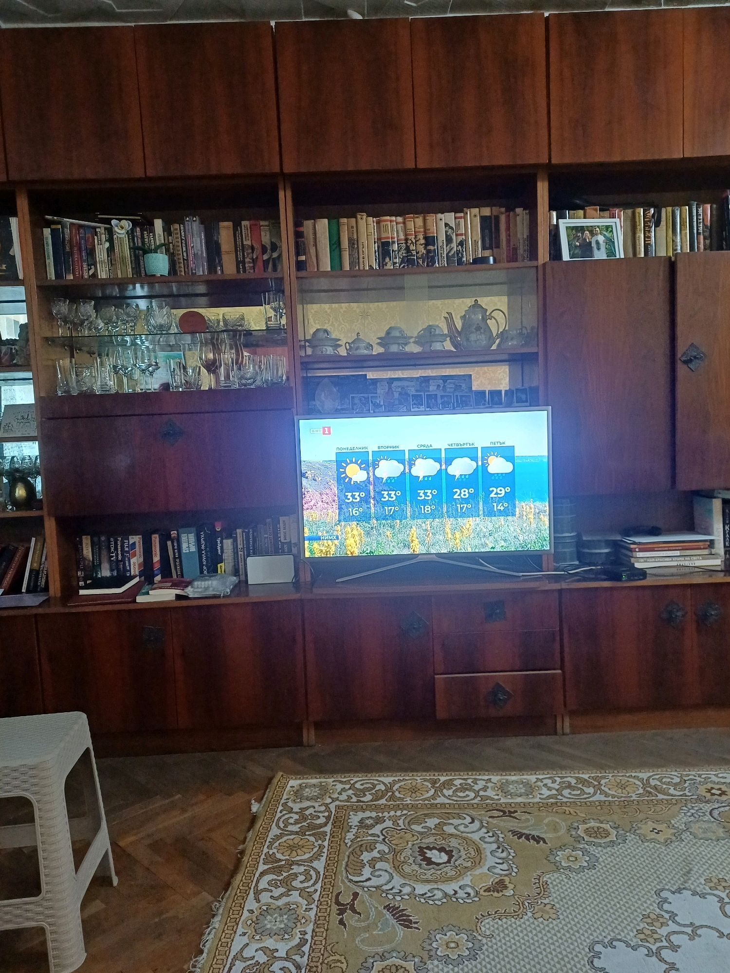 This is a medium-shot, interior photograph taken in a room in the city of Cheshnegirovo, Bulgaria. The room features a large, wooden wall unit filled with books and decorative items, and a television displaying a weather forecast. The wall unit is dark brown wood with glass-fronted shelves and solid doors. The shelves contain various books, glassware, and some decorative teapots. A framed photograph is visible on one shelf. The television, positioned on a lower shelf, shows a weather forecast with icons representing clouds and sun, along with temperature readings in Celsius. The foreground of the image includes a patterned rug and a white plastic stool. The lighting appears to be from overhead fixtures, casting a warm glow on the scene.