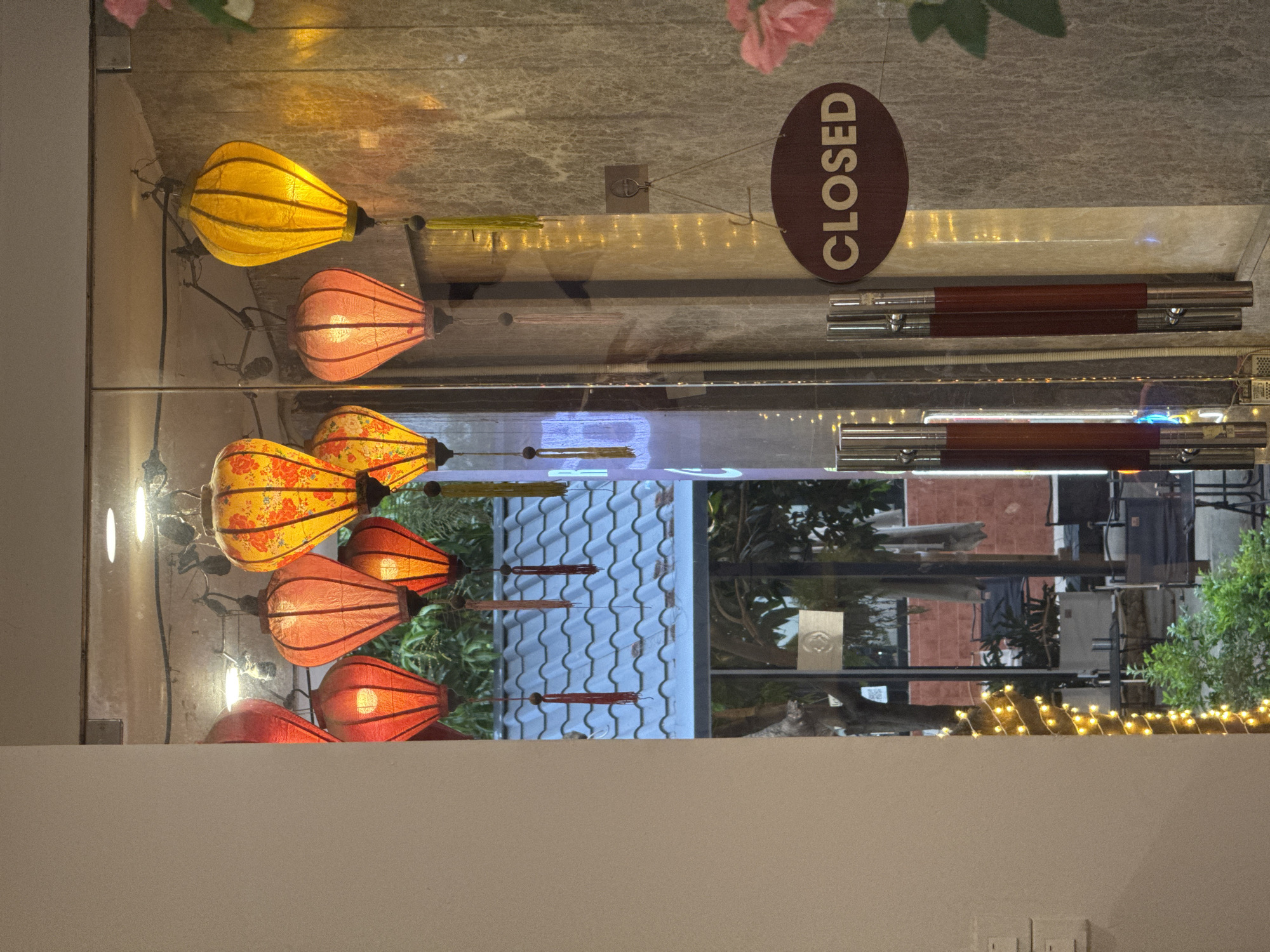 The image shows a display of decorative lanterns, likely in a shop window or restaurant in Hanoi, Vietnam. The lanterns are various shades of yellow, orange, and red, and some are adorned with floral patterns. They are illuminated from within, creating a warm and inviting glow.
The display is seen through a glass pane, which reflects some of the interior. A circular "CLOSED" sign is prominently displayed on the glass, indicating the establishment is not open. In the background, through the glass, architectural elements of a building with a tiled roof can be seen, along with greenery and other decorative lights. The lighting suggests it could be evening or nighttime. The overall scene evokes a sense of traditional Vietnamese decor and ambiance.