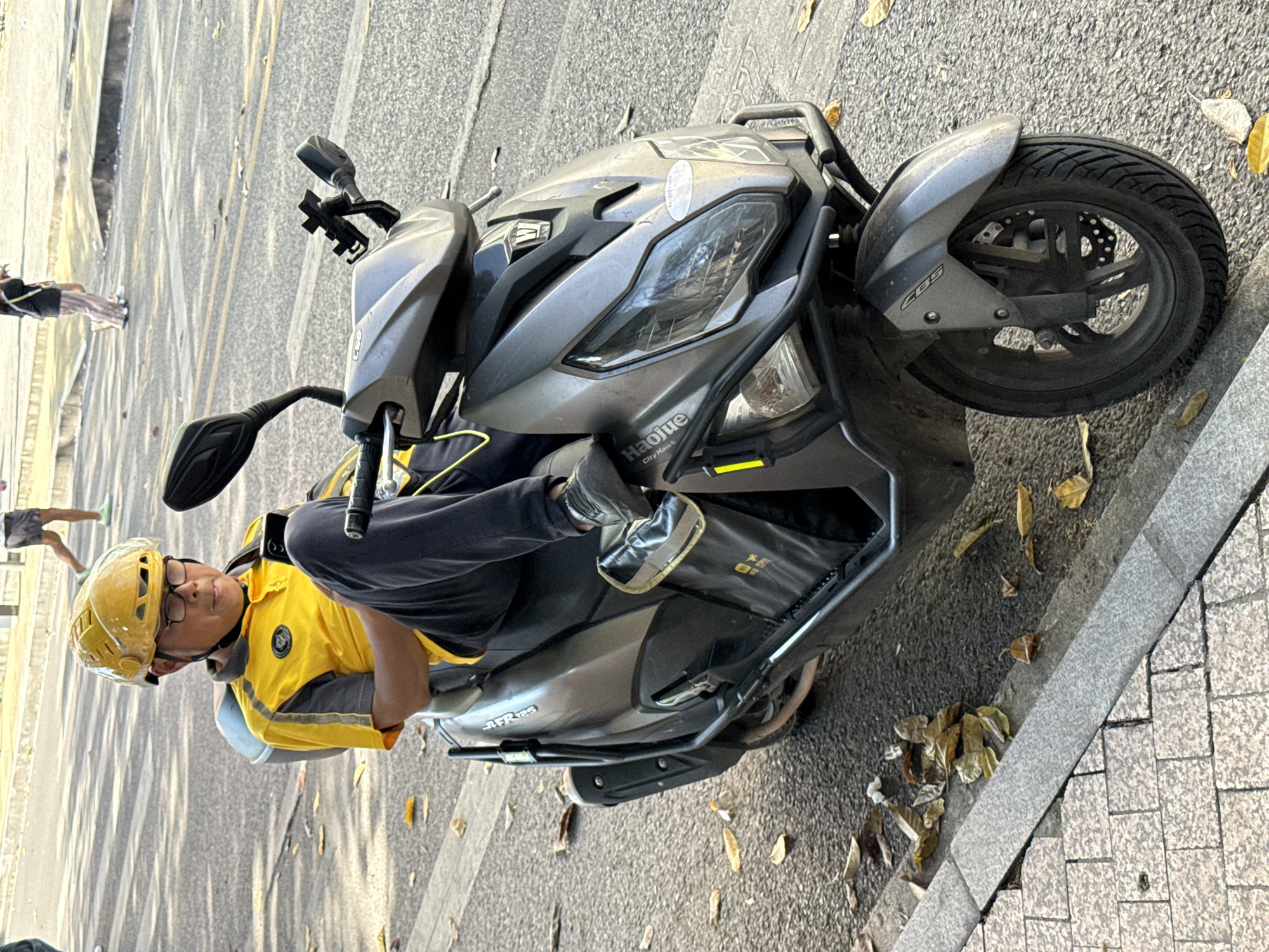 A delivery driver in a yellow shirt and helmet sits astride a grey AFR125 scooter on a street in Jiulongpo District, China. The street has a sidewalk with a curb; the gutter is covered with fallen leaves. The scooter has a brand label that reads "Haojue" and "City Hawk L". A few pedestrians are visible in the background.