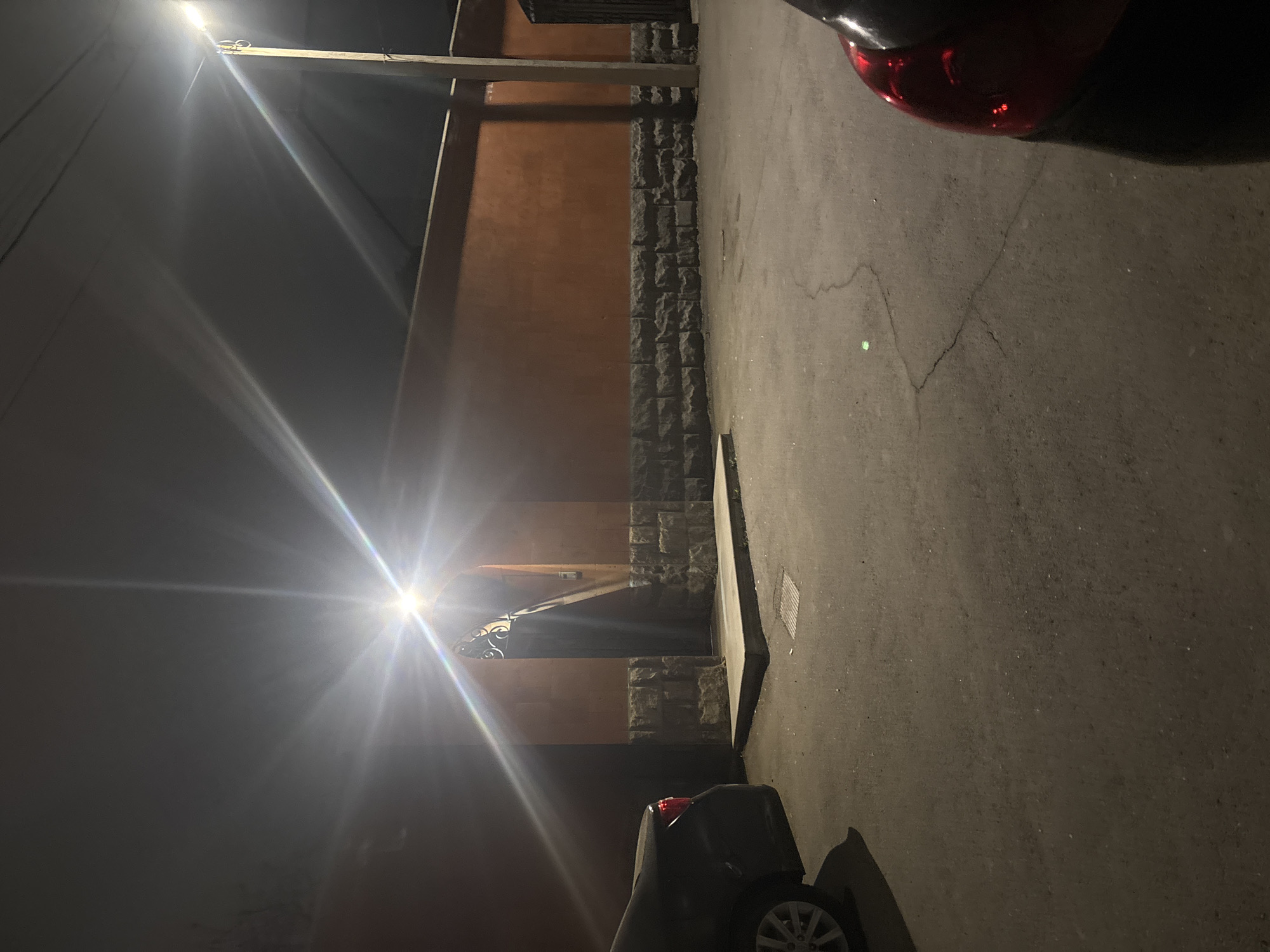 The image depicts a night scene in Yerevan, Armenia, near a building with an orange wall and stone trim. The street is illuminated by a bright street lamp, creating a strong glare. Two cars are partially visible, one with a red taillight in the upper right and the other with a wheel visible in the lower part of the image. A shadowed doorway is located on the left. The ground is paved.