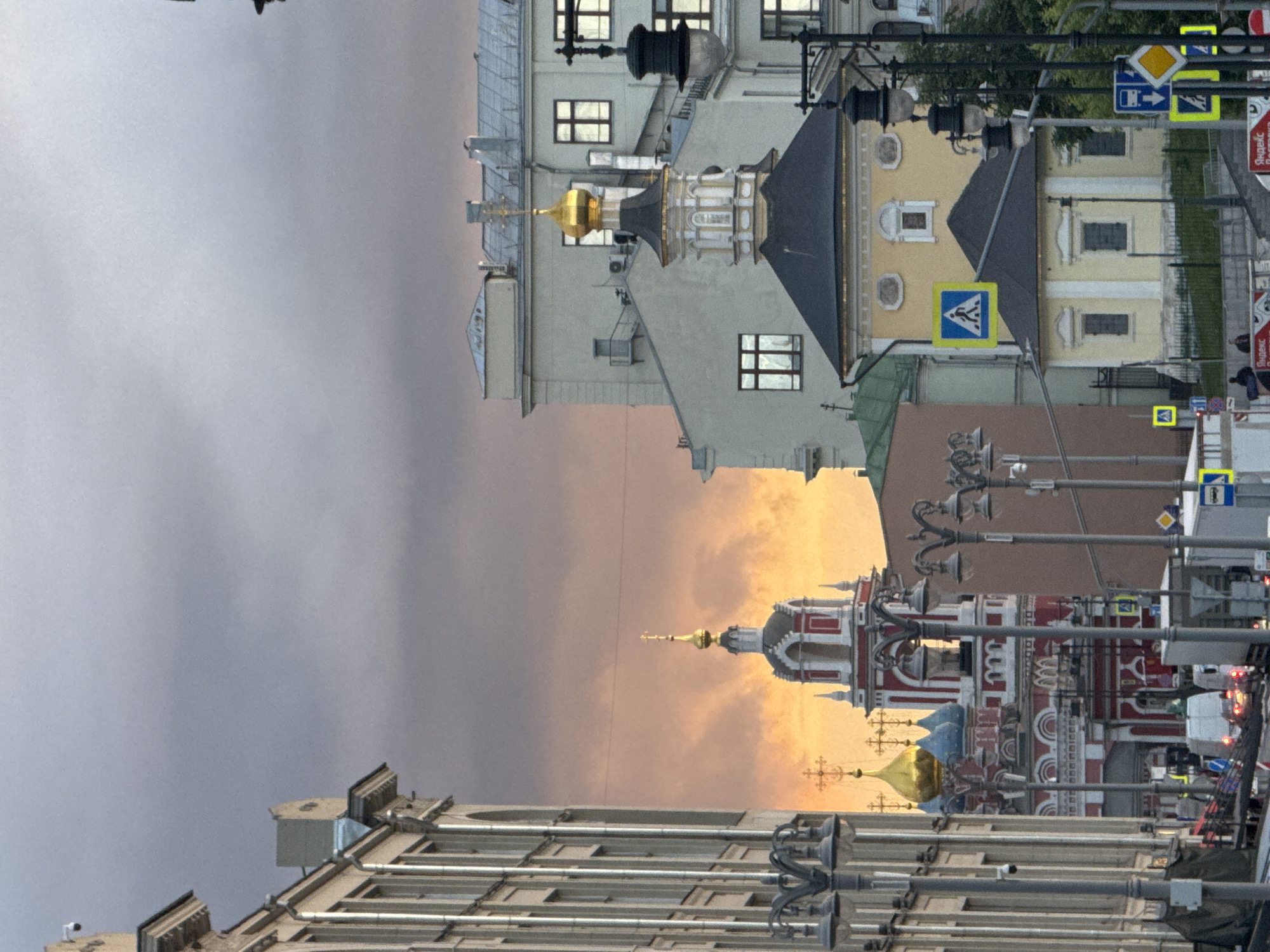 The image depicts a street scene in Moscow, Russia, at dusk or dawn, with the sky displaying orange and gray hues. The architecture of the buildings suggests an old European style, possibly from the 18th or 19th century. Several church towers with golden domes and crosses are visible, typical of Russian Orthodox churches. Streetlights line the road, indicating urban development. Traffic signals and road signs are also present, showing pedestrian crossings and bus stops. Vehicles are likely moving in the street, as inferred from the blurred lights. The overall impression is that of a city with a rich historical and religious heritage.