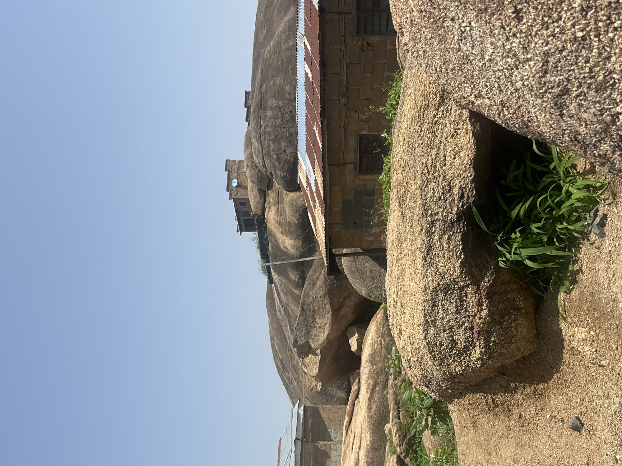 The image captures a landscape in Jos, Nigeria, dominated by large granite rock formations. In the foreground, rough-textured, beige-colored boulders are visible, some partially covered by dry, sandy soil and sparse green vegetation. In the mid-ground, several structures are present. One is a building with walls made of light brown bricks, topped with a rusted corrugated metal roof. Adjacent to it, another building with a light blue corrugated metal roof is partially visible on the left. The background features a massive, rounded granite rock formation. Perched on top of this rock formation are two small, brick structures, one of which has a satellite dish. The sky is clear and pale blue.