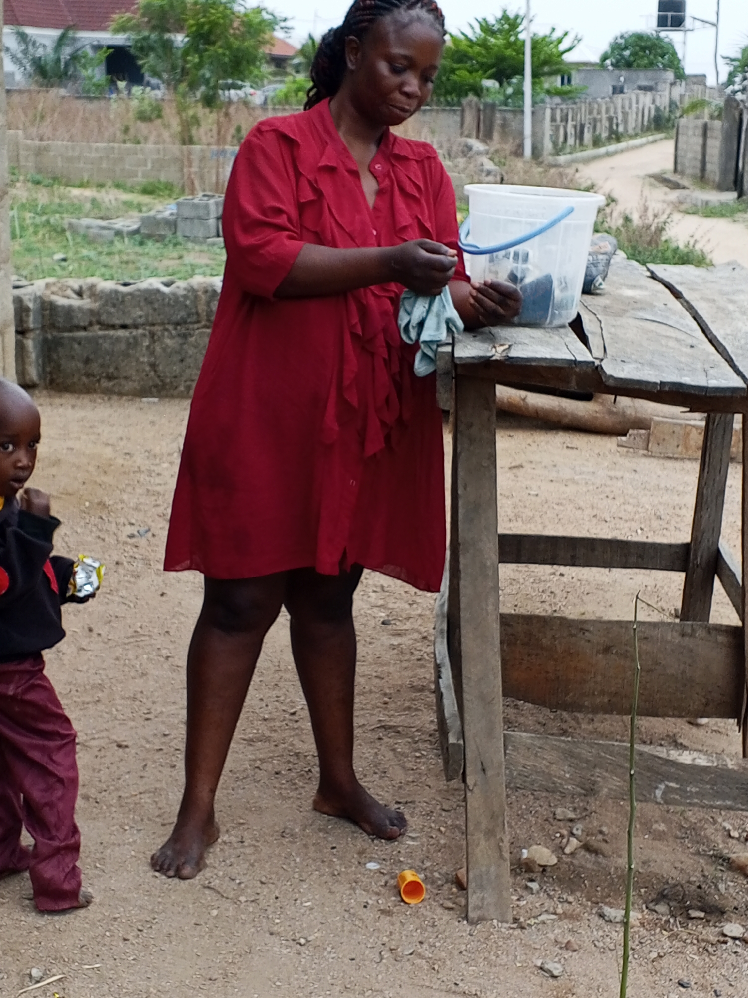 In Adonkolo, Nigeria, a woman with braided hair, wearing a maroon dress, stands barefoot beside a weathered wooden table, holding a blue cloth, near a clear bucket containing dark objects. A small child in a dark jacket and maroon pants stands to her left, holding a small, shiny package. An orange cap lies on the ground near the table's leg. The surrounding environment includes sandy ground, sparse vegetation, partially constructed brick structures, and a dirt road.