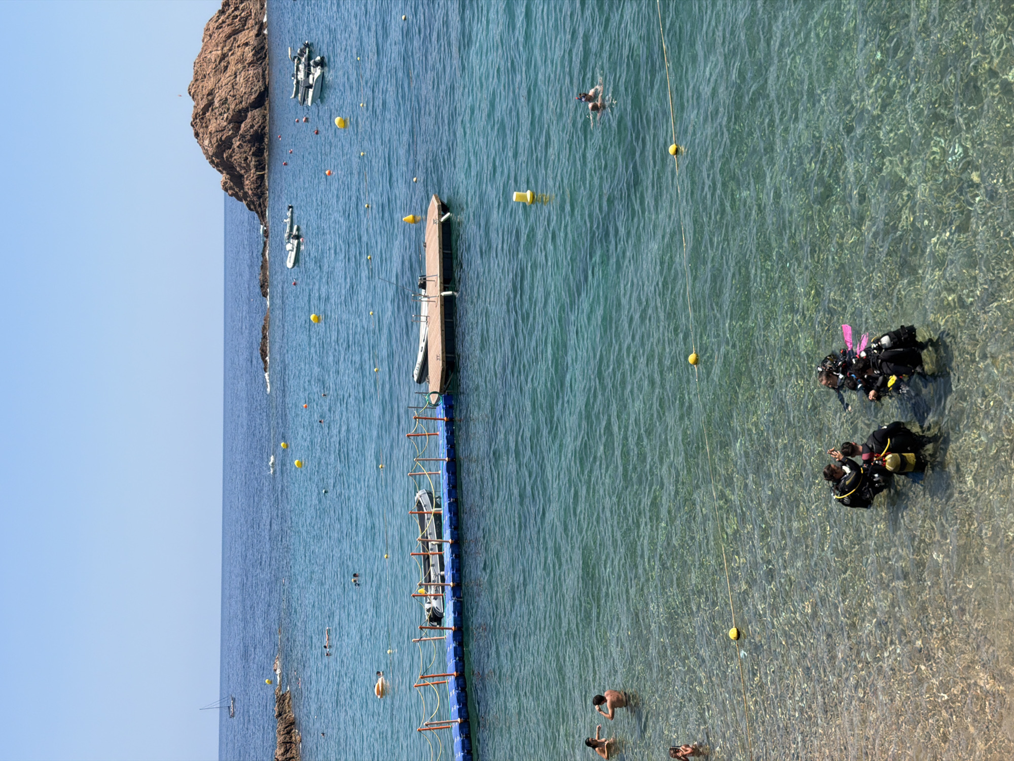 The image depicts a sunny day at a Mediterranean beach, likely in Tossa de Mar, Spain, given the rocky coastline and clear turquoise waters characteristic of the Costa Brava. The foreground shows a group of four individuals in scuba gear, standing in shallow, clear water near the sandy seabed. They appear to be preparing for or concluding a dive, with one person adjusting equipment.
Moving towards the middle ground, the water becomes deeper and a blue floating walkway with orange netting stretches across it. Several small boats are moored or docked along the walkway, including inflatable dinghies and a larger wooden platform with a small boat attached. In the distance, a large, rugged rock formation rises from the sea, a common sight along the Spanish coast.
The water is dotted with yellow buoys, marking an area. Further out, several people are swimming or relaxing in the water. A lone sailboat is visible on the horizon against a clear blue sky. The overall scene conveys a sense of leisure and aquatic activity.