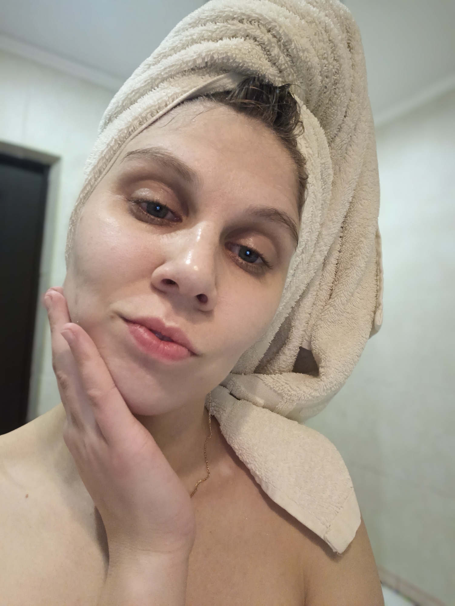 A medium-close-up selfie captures a fair-skinned woman, likely in her 20s or 30s, looking directly at the camera. Her dark hair is wrapped in a light-colored, fluffy terry cloth towel, suggesting she has recently showered or is engaging in a skincare routine. Her skin appears dewy and hydrated, with a subtle sheen visible on her forehead, nose, and cheeks. She gently touches her right cheek with her right hand, her fingers spread, which could indicate a pose or that she is feeling her skin after application of a product. Her lips are slightly parted in a soft pout, and her expression is neutral yet engaging.
Her eyes are a prominent feature, appearing dark, and in some close-ups, there's a slight reddish tint around the lower lash line, possibly from recent washing or a skincare product. Her eyebrows are naturally shaped and well-defined. A thin, delicate gold chain necklace is visible on her collarbone and shoulder.
The background is simple and out of focus, consisting of light-colored walls, with a dark doorway or frame partially visible on the left side, consistent with an indoor setting like a bathroom. The overall impression is one of freshness and post-shower relaxation or a beauty regimen. No text is visible in the image.