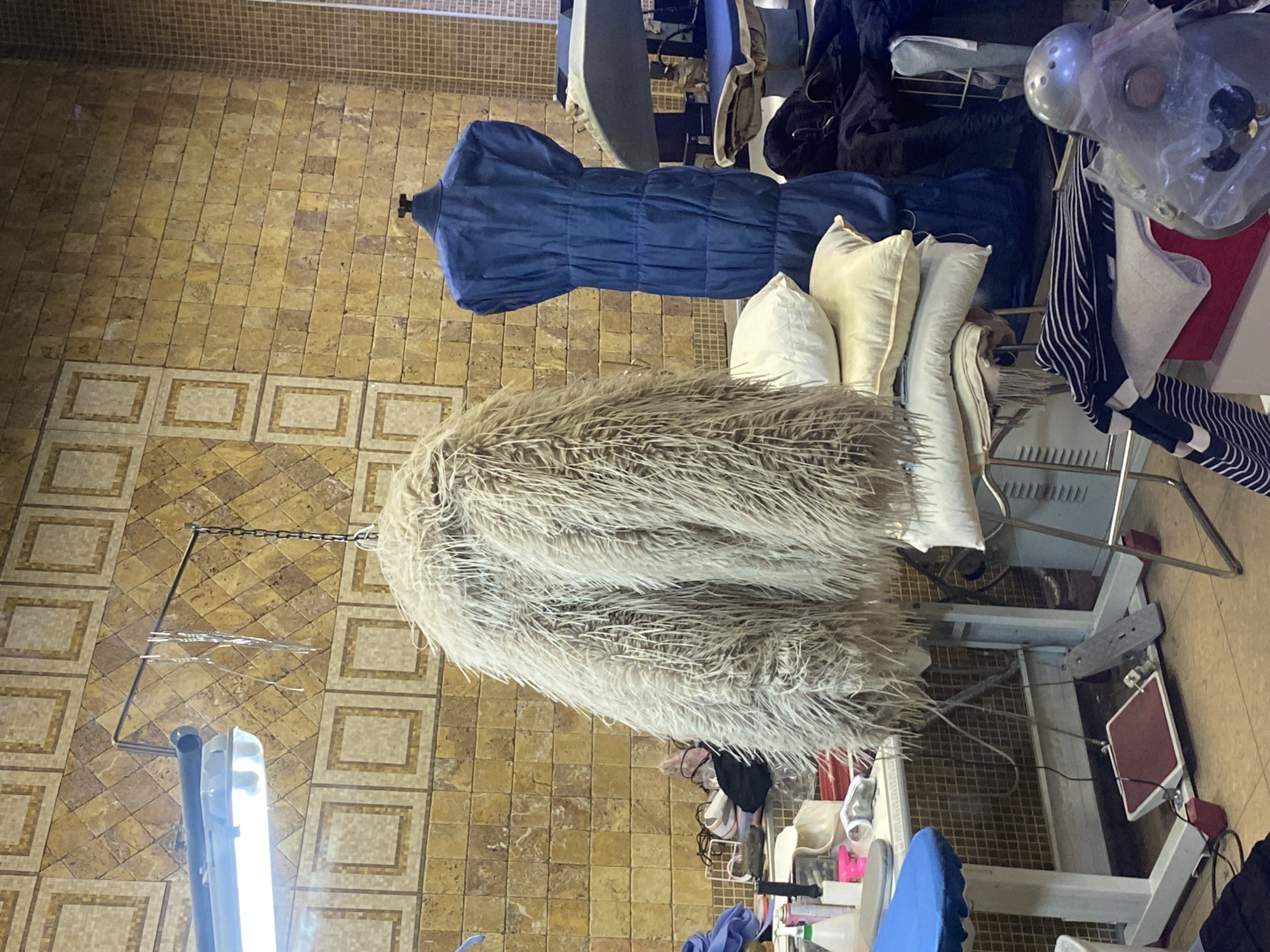 The image depicts an indoor scene, likely a textile workshop or studio, presented ninety degrees counter-clockwise. In the foreground and center, a large, shaggy, light-colored object, possibly a faux fur garment or fabric piece, hangs from a metal chain. To its right, a blue, pleated garment is displayed on a headless mannequin torso.
Behind the mannequin and the hanging fur, several light-colored pillows or cushions are stacked on a metal rack or shelf. The upper right and background areas are cluttered with various items, including dark fabrics, bags, and components of machinery or tools, such as a silver-colored metallic object. A black and white striped fabric is also visible on a rack.
The floor is covered with light brown, square tiles, some featuring decorative patterns, while a section of a wall with smaller, mosaic-like tiles is visible in the top left corner (original image top). A bright fluorescent light fixture is prominent in the immediate foreground. Various pieces of equipment, including metal stands, racks, and a white structure with a red rectangular foot pedal, are present throughout the space. No people are visible, and there is no discernible text. The scene suggests an active, functional, and somewhat cluttered workspace, likely situated in Moscow, Russia.