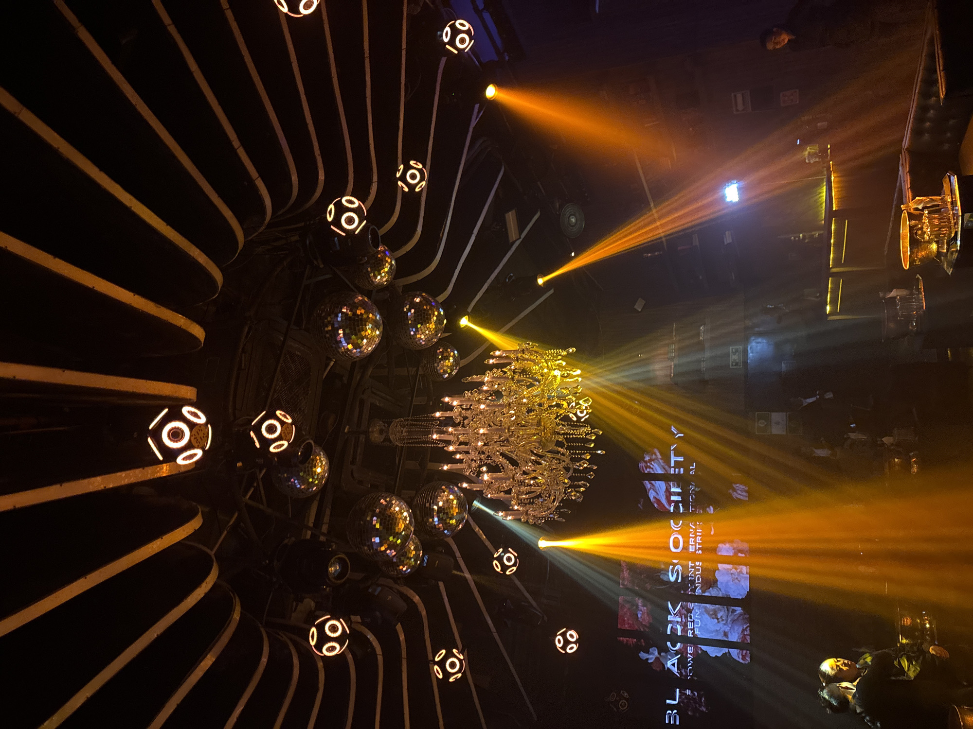 The image depicts the interior of a bar/club environment, likely in Puebla, Mexico. The space features a decorative ceiling with curved, parallel panels interspersed with disco balls and illuminated spherical lights. A large chandelier hangs centrally. Spotlights project beams of yellow/orange and white light across the space, illuminating a back wall with the words "BLACK SOCIETY INTERNATIONAL". People are visible in the background, possibly patrons and staff. The environment has a dark and atmospheric ambience.