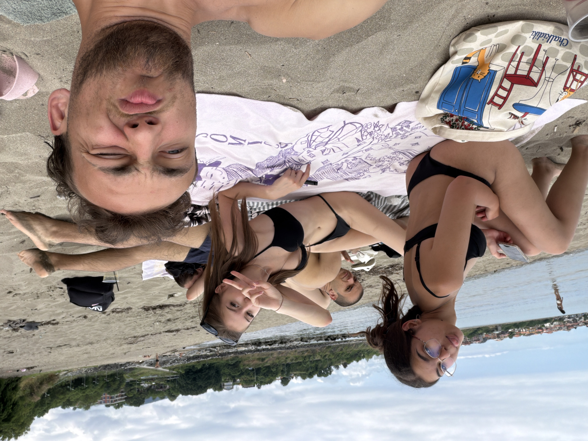 The image captures a group of young people at a beach in Tsarevo, Bulgaria. In the foreground, a young man with a beard and messy hair is making a funny face directly at the camera. To his left, a young woman with glasses is seated on the sand, holding a smartphone and sticking out her tongue. Behind her, another young woman in a black bikini is lying on a patterned towel, striking a peace sign with her hand. Two young men are also visible, lying on the sand behind the women. The background shows the sea, a coastline with buildings, and lush green hills under a cloudy sky. A printed tote bag with cartoon cats and furniture is placed on the sand next to the seated woman.