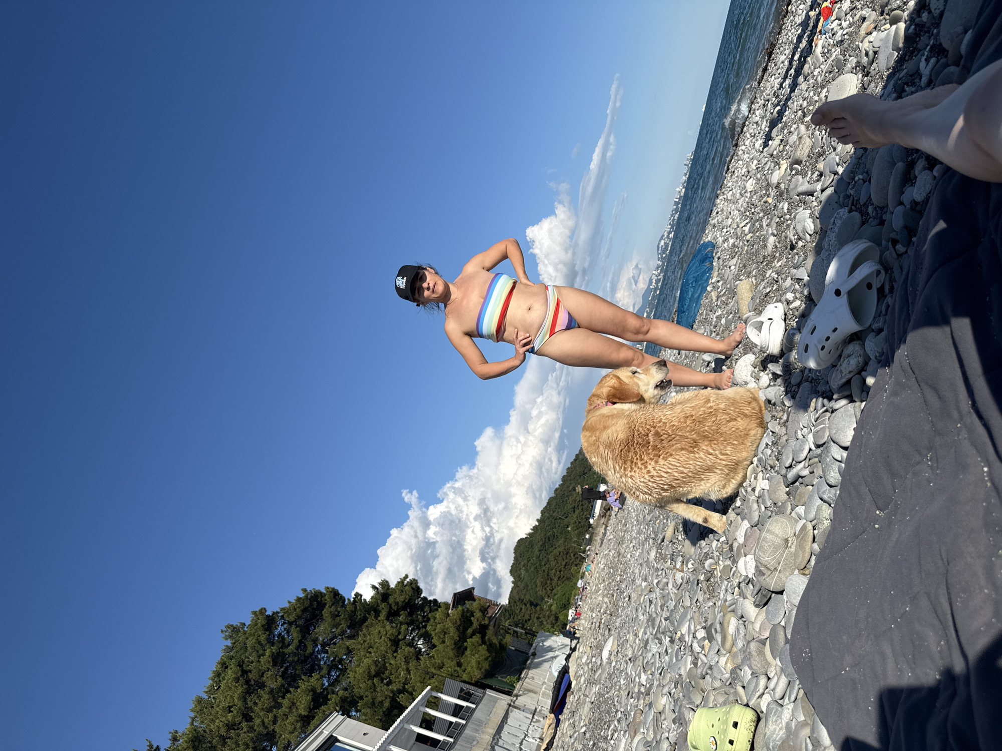 The image depicts a sunny day at a beach in Sochi, Russia. A woman in a rainbow-striped bikini and a black baseball cap stands on a pebble beach with her hands on her hips, posing for the camera. Beside her, a light brown dog is lying down, looking away from the camera. The sea is visible in the background, with gentle waves and a hazy distant coastline. The sky is a clear, bright blue with a few fluffy white clouds. Trees and some buildings are visible on the hillside behind the beach. White Crocs are scattered on the pebbles, along with a blue tarp and other beach accessories. A person's legs and feet are visible in the foreground, suggesting they are sitting or lying on the beach. The overall mood is relaxed and summery. The time of day appears to be midday, given the bright sunlight.