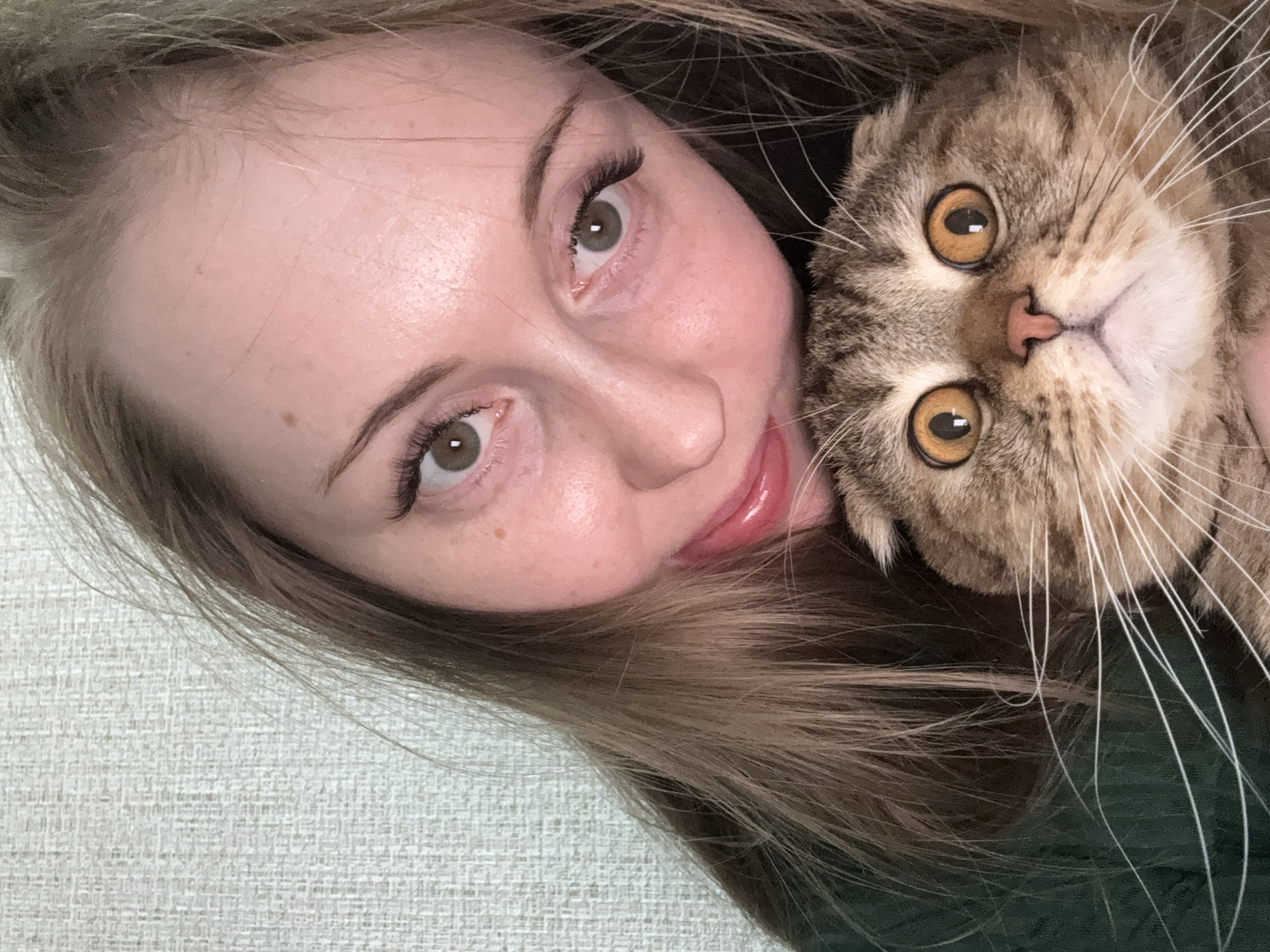 A close-up, diagonally oriented portrait features a young woman and a tabby cat, both looking directly at the camera. The woman's face occupies the left side of the frame, showing her forehead, eyes, nose, and mouth. She has fair skin, light brown hair that cascades around her and behind the cat, and striking green/hazel eyes with long, dark eyelashes. Her eyebrows are neatly shaped, and her lips are a glossy, soft pink, hinting at a slight, pleasant smile.
Snuggled against the woman's head on the right is a cat, possibly a Scottish Fold or British Shorthair mix, identifiable by its round face and potentially folded ears (though not fully visible). The cat has large, luminous amber-orange eyes that are wide and alert, a small pink nose, and prominent white whiskers. Its fur is a mix of light and dark brown stripes. Both subjects appear engaged with the camera, creating an intimate and endearing moment.
The background is a textured, light-colored surface, possibly an off-white or light gray fabric, visible in the lower left corner. The woman's hair and a dark green garment, likely her clothing, fill the remaining background space around the cat. The lighting is soft and even, suggesting an indoor setting. There are no discernible location cues, specific time of day, or visible text. The overall mood is affectionate and serene.