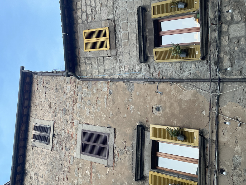This image depicts a section of two adjacent multi-story buildings in Castel del Piano, Italy, under an overcast sky. The left building features a facade of mottled brown-grey plaster and exposed stone, with areas of visible red brick. The right building exhibits a facade constructed predominantly from grey stone blocks. A dark brown vertical downspout runs along the seam between the two structures.
Five windows are visible across both facades. On the left building, the uppermost window has weathered grey-brown closed louvered shutters. Below it, a second window features dark brown closed louvered shutters. The lowest window on the left building has open yellow-green louvered shutters, revealing a multi-pane glass window. Two small green plants in grey metal buckets are suspended from the lower hinges of these open shutters.
On the right building, the uppermost visible window displays bright yellow closed louvered shutters. The lower window on this building also has open yellow-green louvered shutters, revealing a multi-pane glass window similar to its counterpart on the left. Two small green plants in grey metal buckets are likewise suspended from the lower hinges of these open shutters.
Multiple dark electrical wires and cables crisscross horizontally across the lower portion of the scene, strung below the two open windows. Several white, bare incandescent light bulbs are attached to these horizontal wires, hanging downwards. A segment of a blue wire runs vertically down the stone wall of the right building, parallel to the dark downspout. A circular vent opening and a metal conduit arm are present on the plaster wall of the left building, between the middle and lower windows. No individuals are present in the scene.