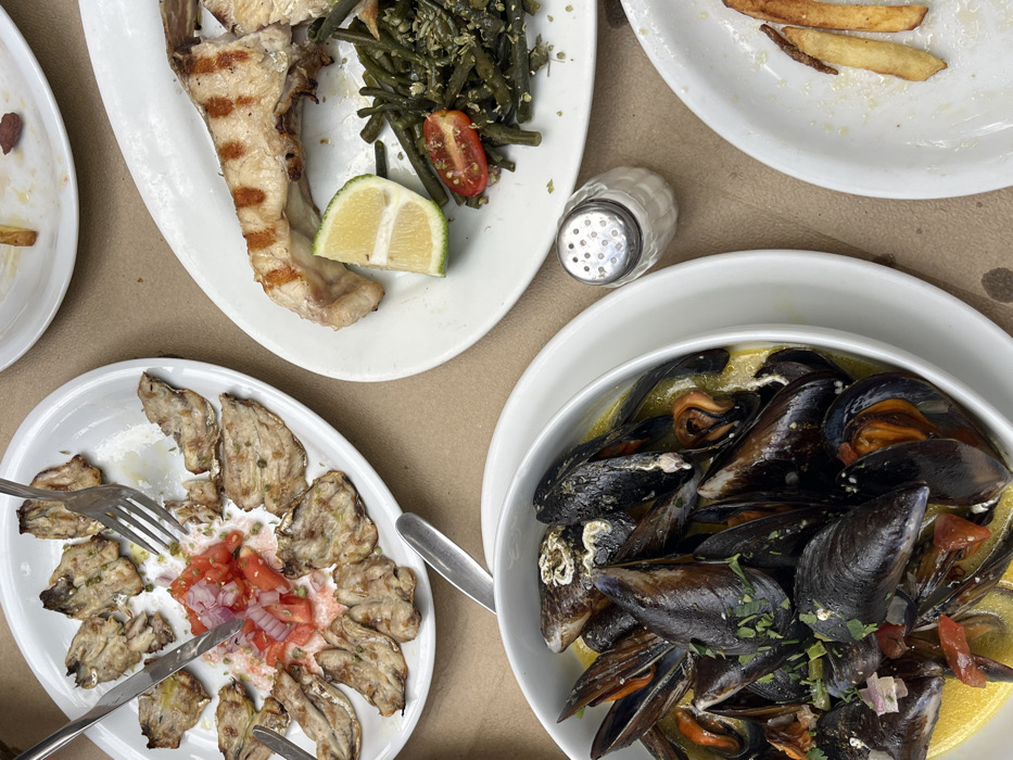 The image displays a dining table scene in Skala, Greece, presenting several partially consumed dishes. In the upper left, a white oval plate holds numerous small, grilled, descaled fish, possibly sardines or anchovies, arranged around a central mound of diced red tomatoes and red onions. A silver fork and knife are embedded in this central garnish, indicating active consumption. To the immediate right, a separate white oval plate contains a grilled fish portion, identifiable by distinct char marks, accompanied by a quarter-slice of lime, a small cluster of halved cherry tomatoes, and a serving of green beans mixed with a granular seasoning. In the lower left, a white bowl contains steamed mussels in a yellow, possibly buttery, broth, garnished with chopped green herbs and diced red vegetable fragments. A small, clear glass salt shaker with a perforated metal cap is situated between the mussel bowl and the grilled fish plate. A partially visible white plate in the lower right shows remnants of French fries and a single dark red item. The table surface is a plain, light brown or beige color. No individuals are visible, but the presence of cutlery in use and partially eaten food confirms dining is in progress.
