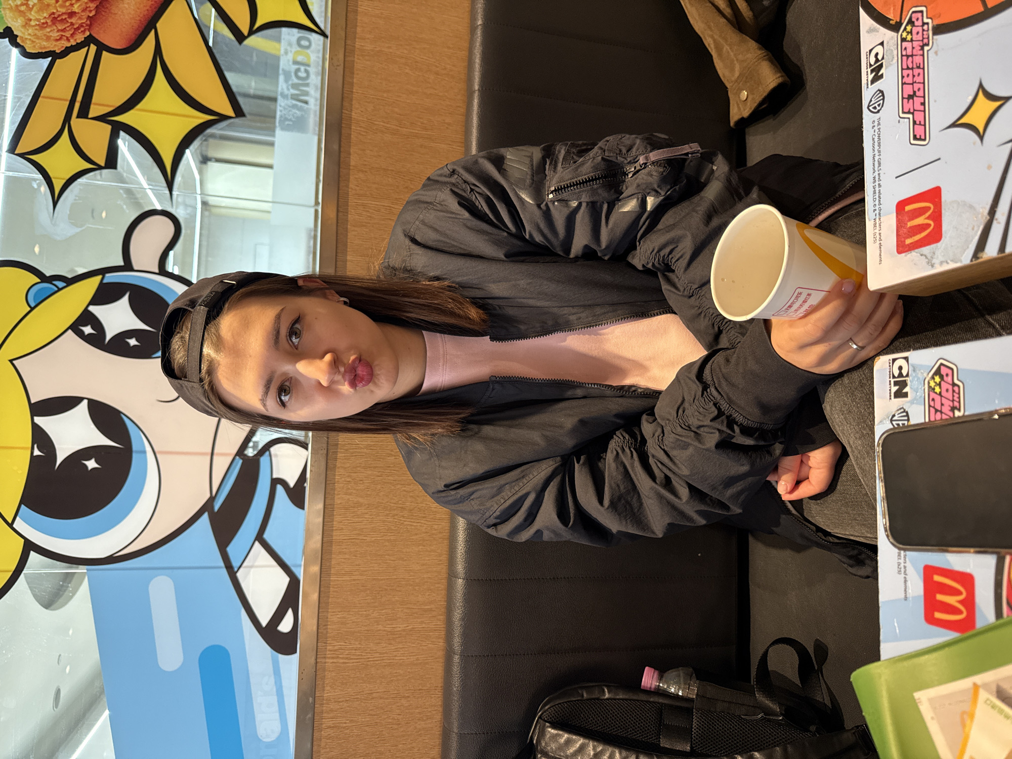 A young woman with brown hair wearing a black cap, pink shirt, and black jacket is sitting at a table inside a McDonald's restaurant. She is holding a partially consumed McDonald's cup and making a kissing face towards the camera. Beside her is a McDonald's box featuring "The Powerpuff Girls" characters. A smartphone is on top of the box. A black backpack is on the floor next to the seating. Through the window behind her, a McDonald's logo and a depiction of "The Powerpuff Girls" are visible. The location is likely in a city with McDonald's restaurants, potentially in Dongcheng District, China based on text appearing on the cup.