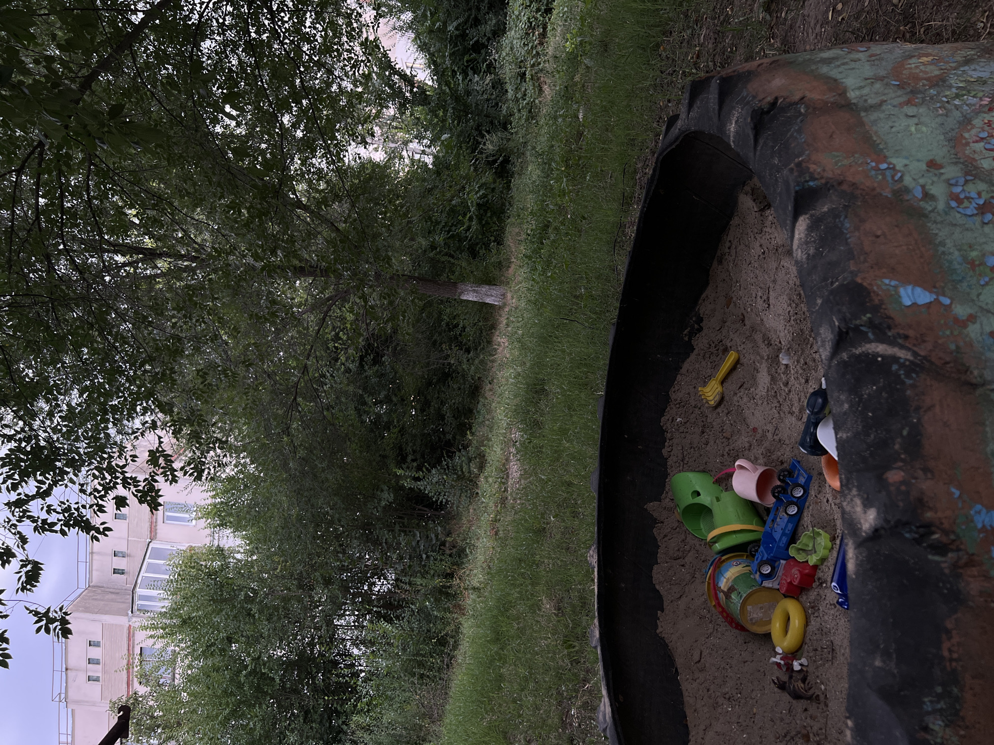 An outdoor scene in Leova, Moldova, features a sandbox made from a repurposed tire filled with sand and various children's toys. The toys include a green plastic sieve, a yellow plastic shovel, a pink plastic cup, a blue toy truck, a yellow ring, and other small colorful plastic items. In the background, a multi-story building with pinkish-beige walls and white windows is visible, partially obscured by dense green trees and foliage. The ground around the sandbox is covered in green grass and some dry dirt. The overall impression is one of an urban or suburban setting with elements of play and nature.