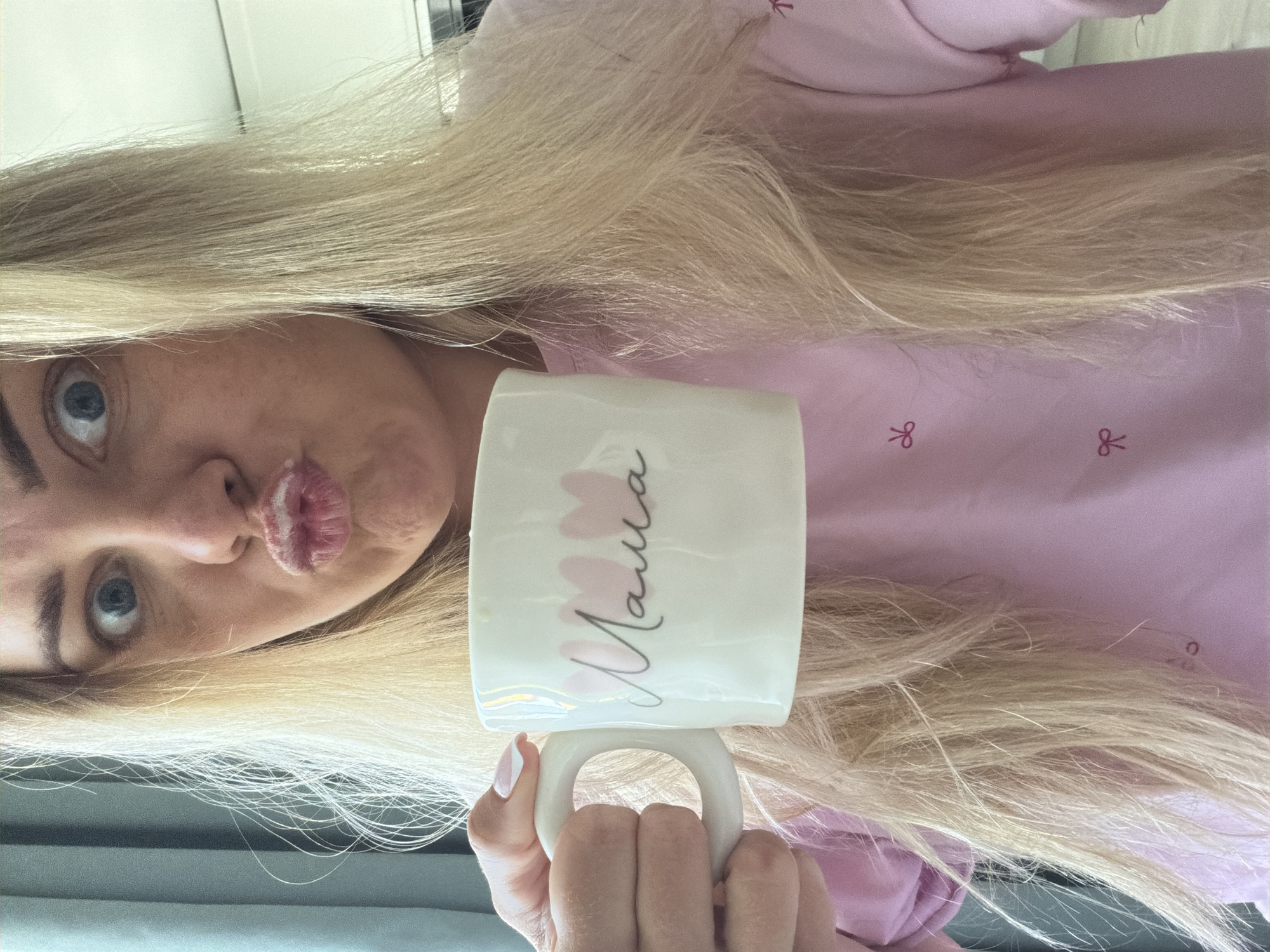 A woman with blonde hair is holding a white mug with the word "Mama" written on it in cursive with pink floral accents. She has blue eyes and is making a kissing face. She is wearing a pink long-sleeved shirt with small bow designs on it. The background appears to be indoors, possibly a home environment. The exact location in Votkinsk, Russia cannot be determined from the image alone.