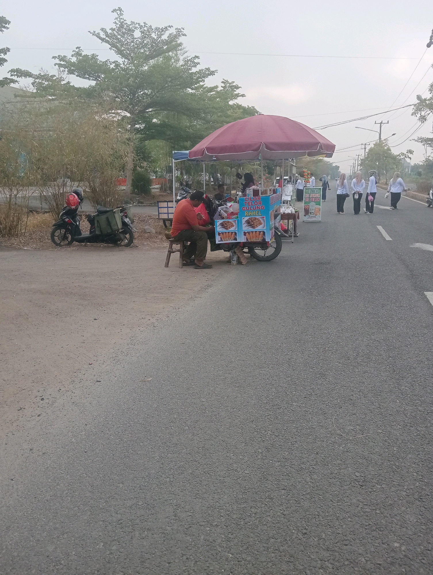 The image depicts a street scene in Sidoharjo, Indonesia, likely in the morning or late afternoon given the soft light and overcast sky. A food cart selling "Cilor Telor Gulung" and drinks like "Es Teh, Es Jeruk, Es Teler" is parked on the side of the road. A man in an orange shirt and dark pants is seated on a wooden stool at the cart, appearing to be working or waiting for customers. Next to the cart, a black motorcycle with a green storage box is parked. In the background, a group of women wearing white tops and headscarves are walking along the right side of the road. The environment includes trees and vegetation on the left side of the road, and a paved road with a white lane marker. Utility poles and wires are visible in the distance.