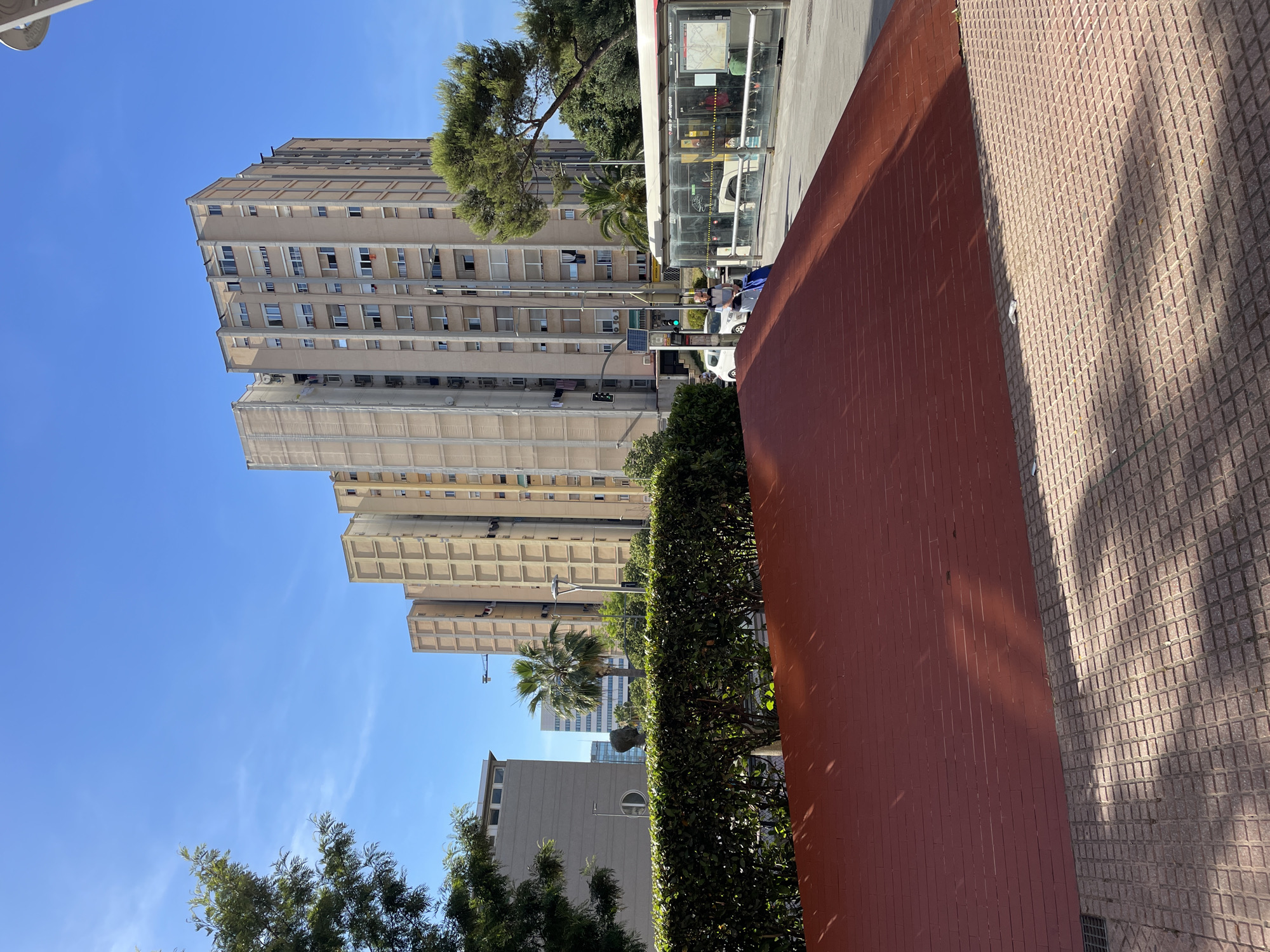 The image depicts an urban scene in Cornellà de Llobregat, Spain, characterized by apartment buildings, a bus stop, and a park area. In the foreground, a red, sloped surface, possibly a decorative element or part of a park structure, is visible, covered in tile on the right side. Beyond this, a meticulously trimmed green hedge forms a boundary. To the right, a bus stop with a transparent enclosure shows people inside, and a bus is partially visible. The background is dominated by several tall apartment buildings with a repetitive facade structure, typical of mid-20th-century urban architecture. A palm tree and other greenery suggest a Mediterranean climate. A man is seen walking with a blue bag next to the bus stop. The sky is a clear, bright blue with a few wispy clouds. The overall impression is that of a sunny day in a residential urban area.