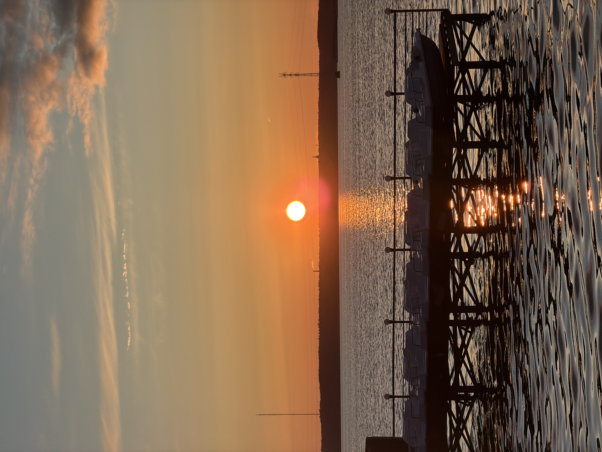 A horizontal photograph captures a tranquil sunset over a large body of water, likely Izhevsk Pond, bathed in warm, golden light. The sun, a luminous orange-red disc, is positioned centrally, low on the horizon. The sky displays a gradient of colors, from soft blue-grey with light clouds in the upper left to vibrant oranges and yellows near the sun.
In the foreground, a dark, silhouetted dock or pier with distinctive X-shaped structural supports extends into the water from the right. Several light-colored, small boats, possibly pedal boats or rowboats, are neatly moored alongside the pier. The water's surface is textured with ripples, reflecting the sun's intense light in shimmering streaks, particularly pronounced closer to the dock.
The distant shore forms a dark, undulating silhouette across the horizon, topped with what appears to be a treeline. On this distant land, a tall structure resembling a radio or transmission tower is clearly visible, along with horizontal power lines stretching across the sky. Another smaller, conical structure, possibly a church steeple, can also be faintly discerned to the left of the main tower. There are no people visible in the image, nor any discernible text. The scene evokes a sense of peace and natural beauty at the close of day.