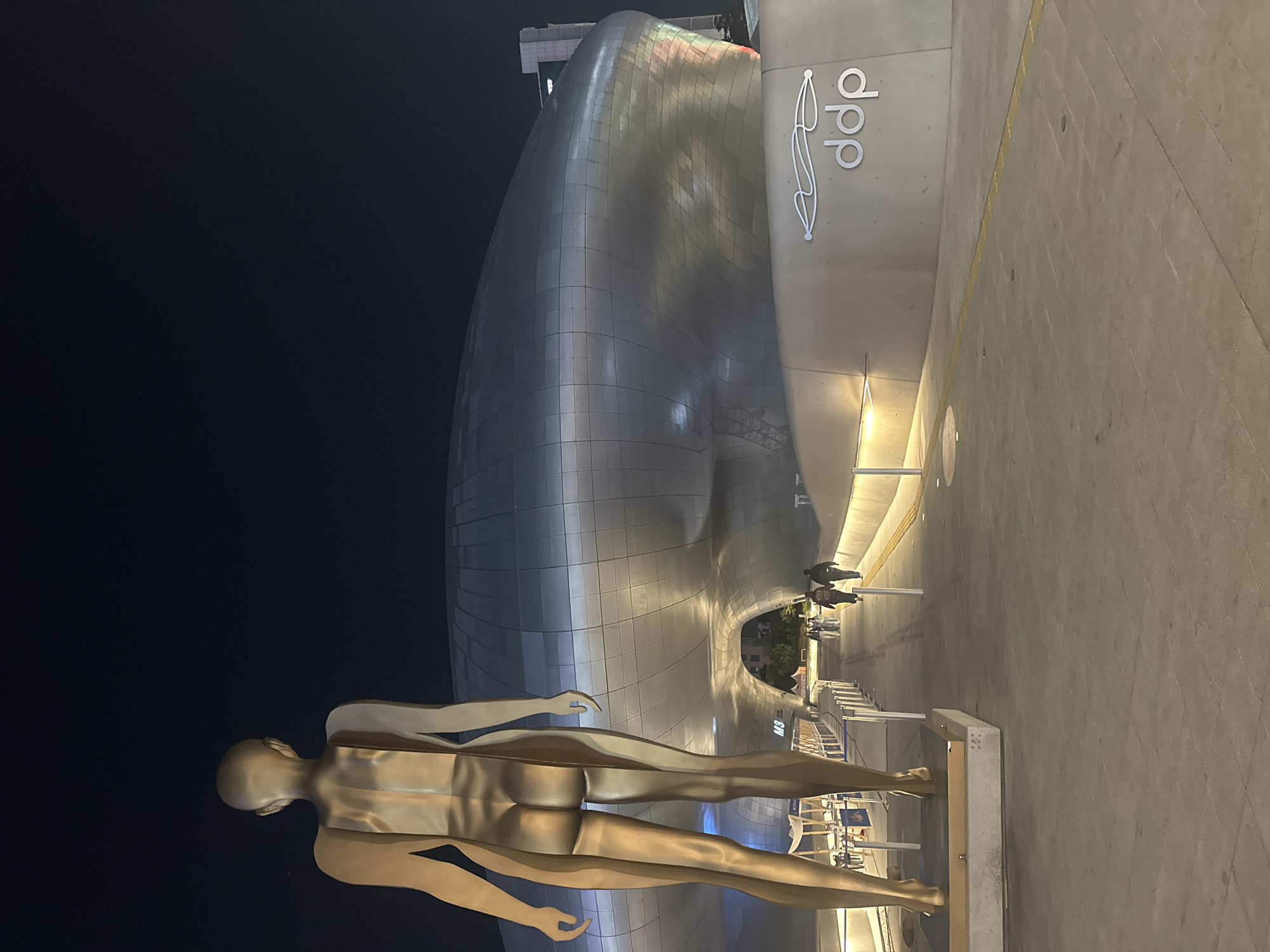 Here's a description of the image:
The photograph shows a nighttime scene in Seoul, South Korea, featuring the Dongdaemun Design Plaza (DDP). The distinctive, futuristic architecture of the DDP is visible in the background, illuminated with artificial lighting. A large, stylized human figure sculpture dominates the foreground, appearing to be gold in color. The sculpture is positioned on a plinth. In the background, a few people are seen walking near the DDP building. The ground is paved and well-lit. The overall environment suggests an urban, modern setting. The DDP logo is clearly visible on the side of the building.