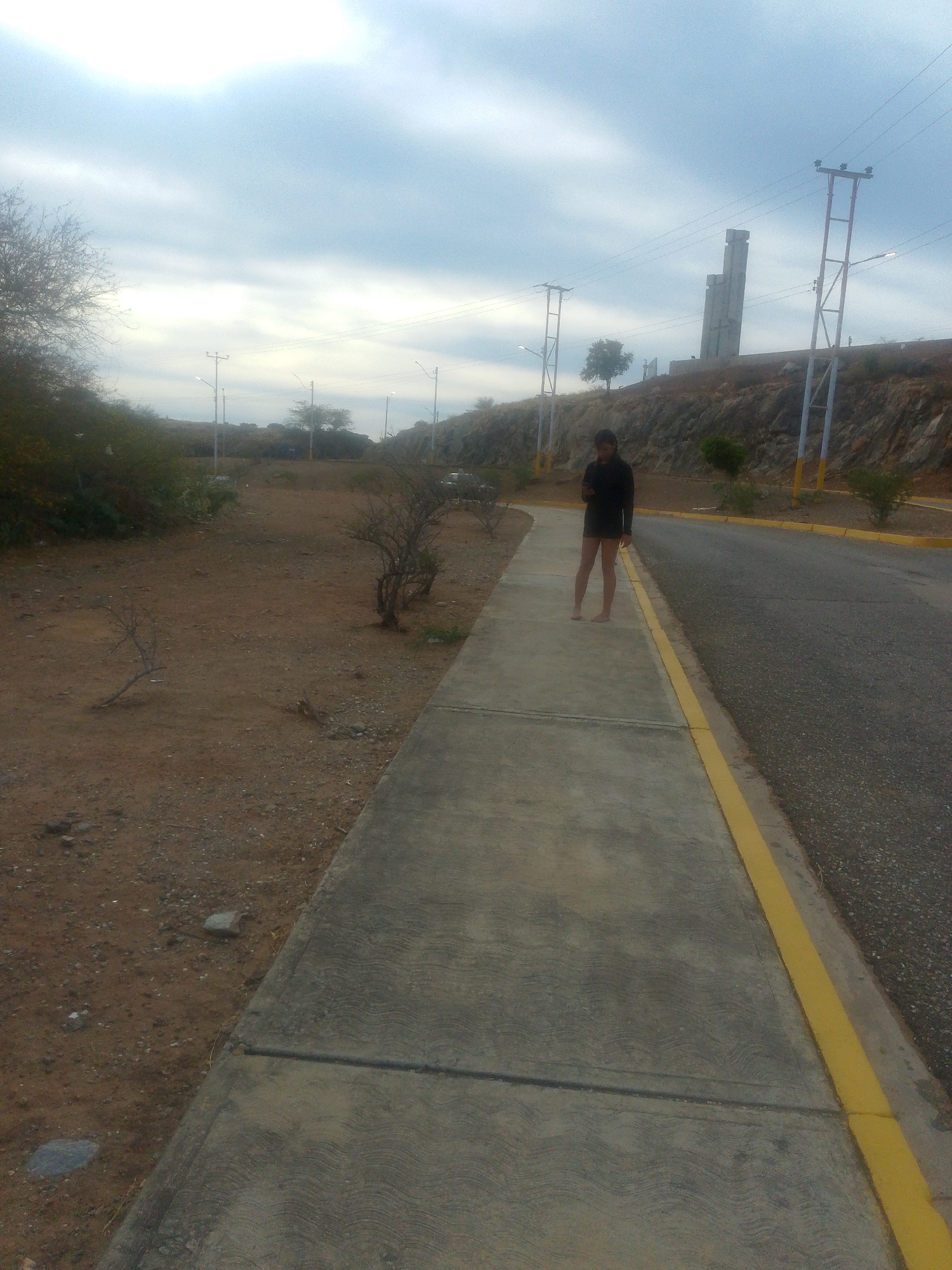 The image depicts a street scene in Juan Griego, Venezuela. A person is standing barefoot on a concrete sidewalk next to a road. The person is wearing a long-sleeved dark-colored shirt or jacket and appears to be looking at a mobile phone. The sidewalk has a yellow painted line along the edge that borders the road. The road surface is dark and appears to be asphalt. Beyond the sidewalk and road are areas of dry, brown vegetation and dirt. Power lines and utility poles line the road, and in the background, a tall concrete structure can be seen on a hill. The sky is overcast.