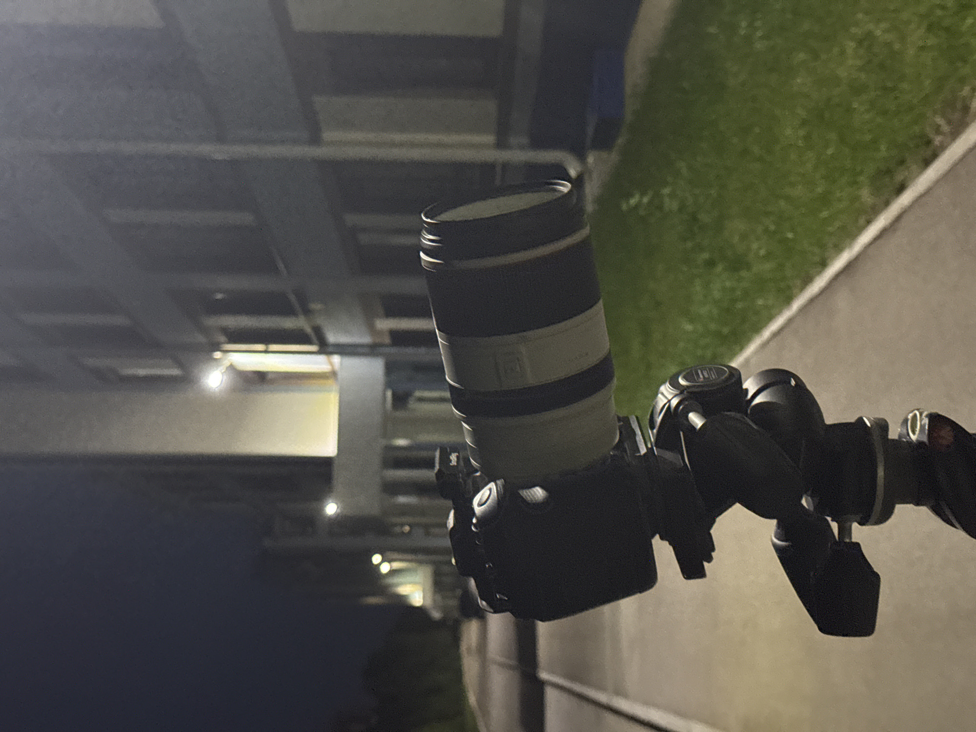 The image shows a camera mounted on a tripod, positioned outdoors at night. The camera lens is white and black. The background includes a building with lit windows and some greenery. The location cannot be determined to be Perm, Russia, from the image alone.