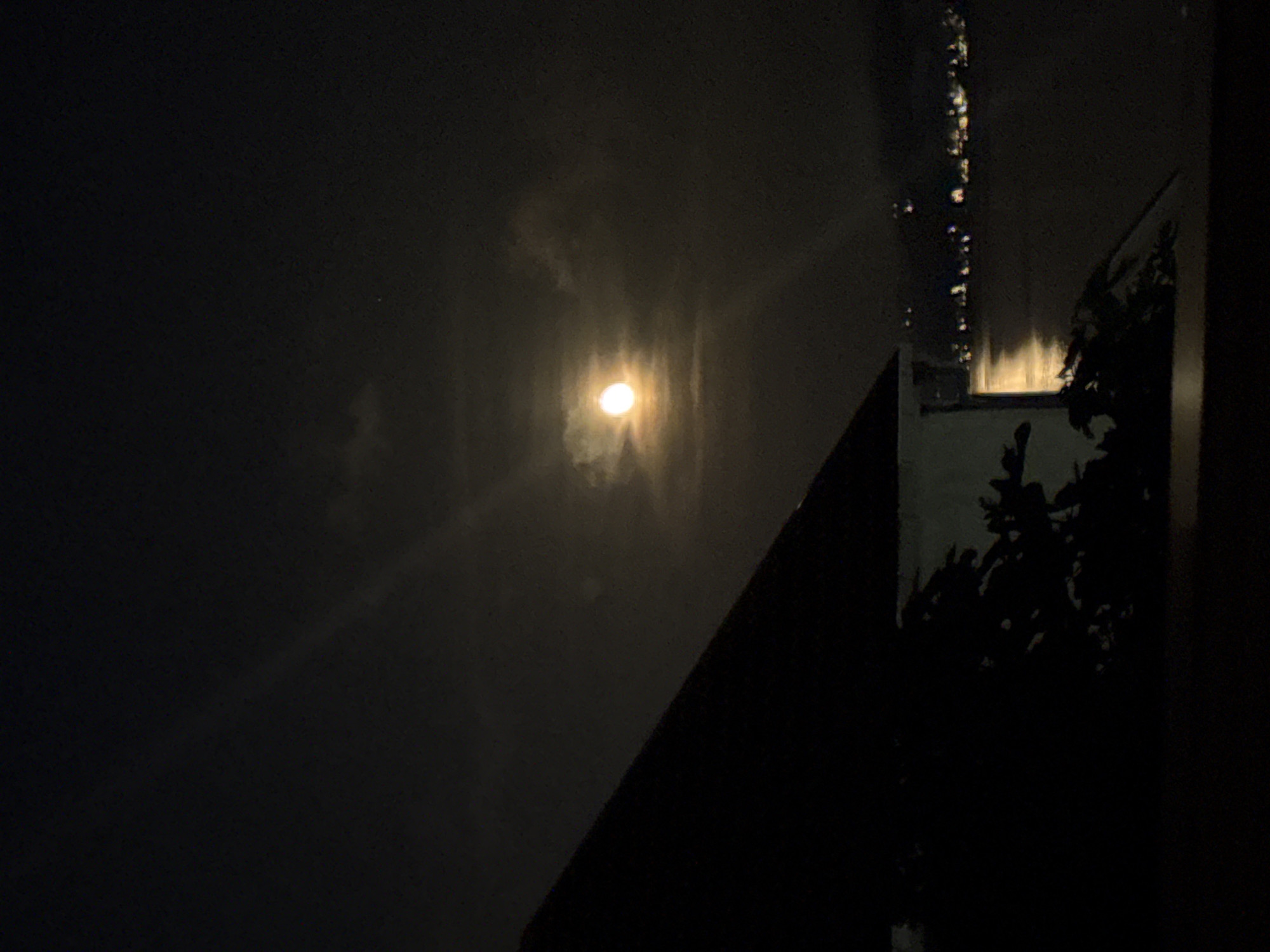 The image presents a nocturnal scene featuring a prominent, brightly glowing moon, partially veiled by clouds or haze, positioned slightly left of the frame's center. The moon's intense light creates a luminous halo and illuminates the surrounding atmospheric conditions.
Silhouetted against the dark night sky, the outlines of architectural structures and foliage are visible on the right side and bottom right of the composition. A distinct dark, triangular shape in the bottom right corner suggests a building's roofline or an adjacent structure. Below this, a cluster of dark, shadowy foliage, likely trees or bushes, adds to the natural elements within the urban or semi-urban setting. On the far right edge, a vertical strip displays several bright, elongated reflections or streaks of light, possibly originating from distant artificial lights or reflections on a surface like a windowpane.
The scene captures a quiet, cloudy night in Bijela, Montenegro, viewed from a vantage point that includes both the sky and parts of the built environment. There is no discernible activity taking place, and no text is visible within the image.
