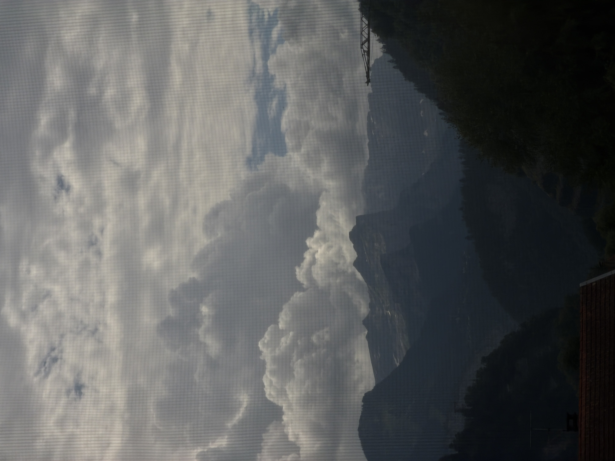 The image depicts a mountainous landscape viewed through a screen, likely a window screen, which creates a faint grid pattern across the scene. The sky is filled with dramatic, billowing clouds, ranging from bright white to dark gray, suggesting an overcast or stormy day. Below the clouds, silhouetted mountains rise, their dark forms contrasting with the lighter sky. The peaks are jagged and appear to be covered in snow in some areas. The lower foreground shows a dark, forested hillside and a portion of a reddish-brown tiled roof, indicating the presence of human habitation. To the right, a construction crane is visible against the sky, its skeletal structure outlined. The overall mood is somber and dramatic, with a sense of the power of nature. The specific location within Cluses, France is not discernible from the image itself; the image depicts a generalized mountainous scene.