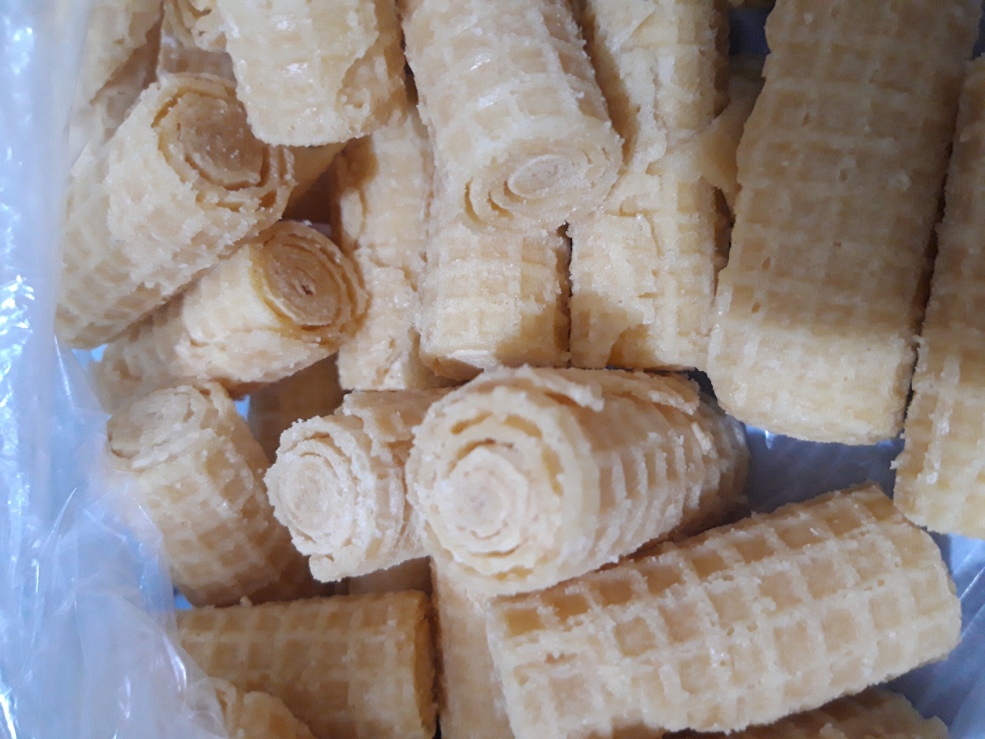 A close-up, overhead shot showcases a plastic bag filled with a pile of wafer rolls. The wafer rolls are light golden brown in color and have a textured, grid-like pattern on their outer surface. Some of the rolls are intact and cylindrical, while others appear to have been broken or are partially obscured. The rolls are stacked and jumbled together, with some facing vertically and others horizontally. The plastic bag is partially visible at the top of the frame, suggesting the treats are being stored or transported. The lighting is soft and diffused, highlighting the dry, crumbly texture of the wafers. The scene is devoid of any human presence or specific environmental context, focusing solely on the food items. The location is unspecified beyond being in Макаров ауылдық округі, Kazakhstan.