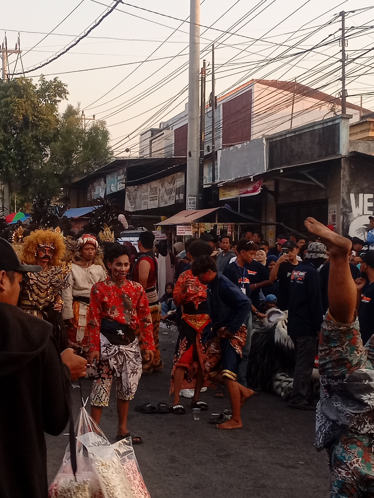 In the city of Margoyoso, Indonesia, a lively street scene unfolds. A group of people, some in traditional attire, are gathered on a paved street lined with buildings. A man with a bright orange, afro-like wig and elaborate costume stands to the left, facing forward. Next to him, another man wears a white garment and a headdress with teeth-like protrusions. In the foreground, a person in a red patterned shirt and a batik sarong is visible, holding a plastic bag filled with white and pink snacks. Behind them, a crowd watches, with some individuals taking photos. The backdrop features buildings with windows and awnings, and overhead, a complex network of power lines crisscrosses the sky, suggesting an urban environment. The lighting indicates it is likely late afternoon or early evening. The overall atmosphere appears festive, possibly during a cultural event or parade.
