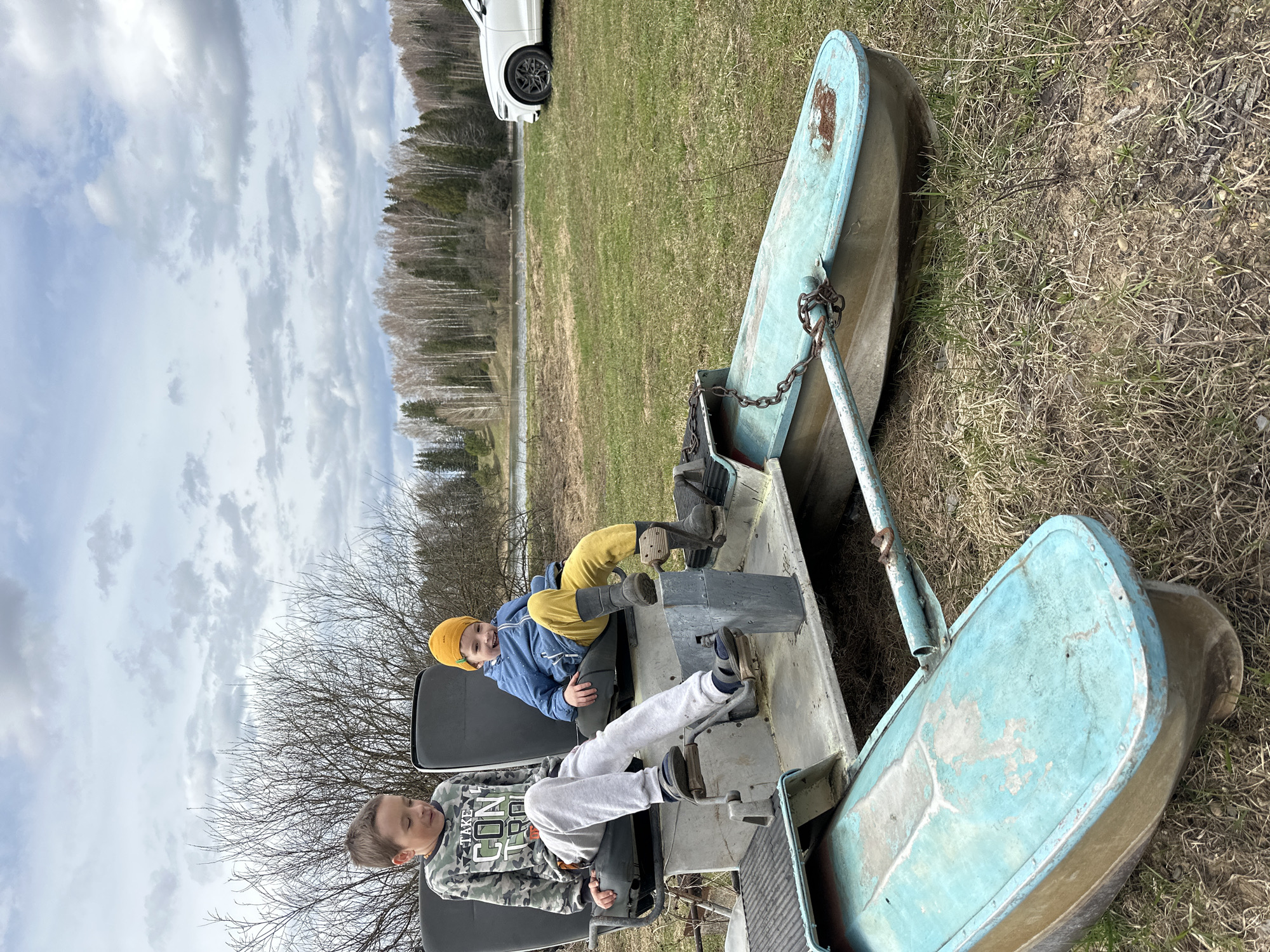 Two boys are seated in a boat-shaped ride in a grassy area, with trees, a body of water, and a white car in the background. The location is Альняш, Russia. The boat ride is comprised of three boat-shaped sections connected together. The boys are sitting on seats with pedal mechanisms. The boy in the foreground is wearing a grey and camo patterned shirt with grey sweatpants, while the boy in the background is wearing a blue jacket, yellow pants, and a yellow hat.