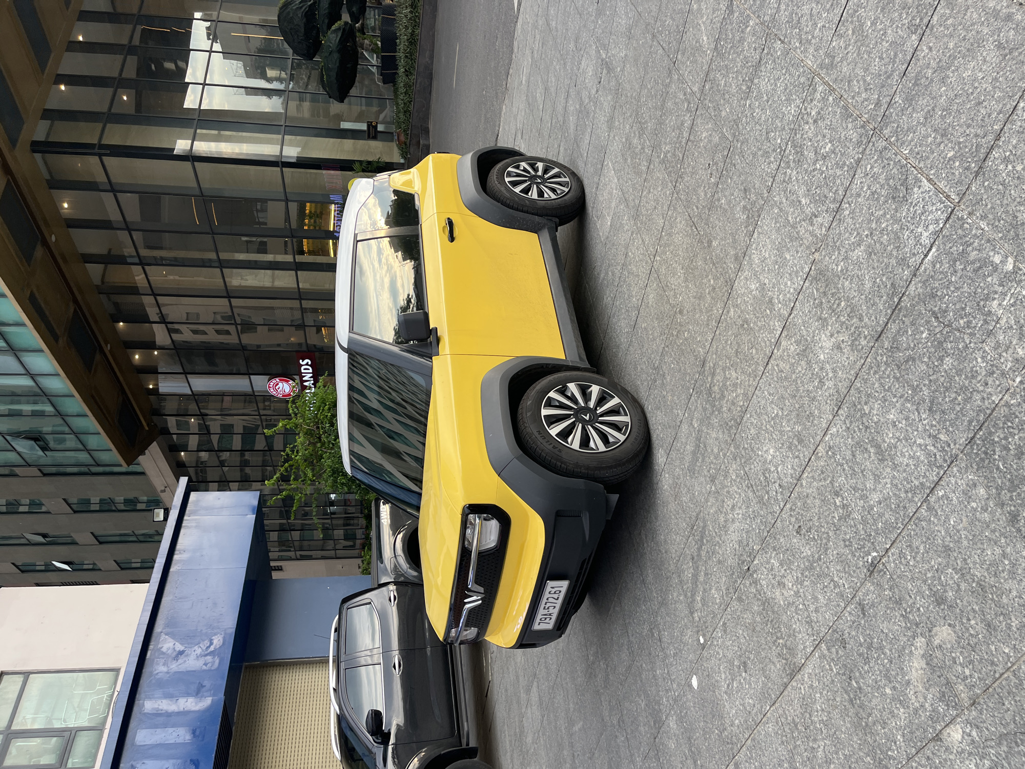 A yellow and white electric car, with license plate "79A-572.61", is parked on a tiled gray surface in front of a building. A dark colored pickup truck is parked adjacent to the car. The building behind has glass windows and reflects the sky. A sign for Highlands Coffee is visible on the building. The location is Bac Nha Trang Ward, Vietnam.