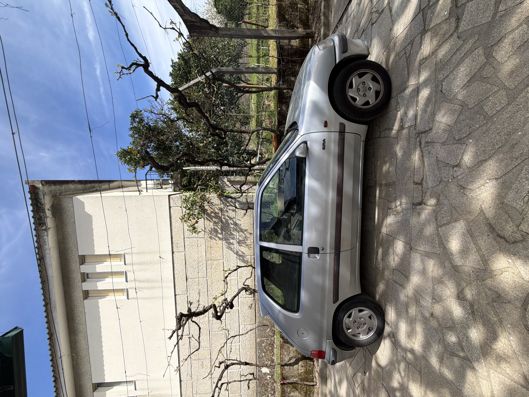 The image shows a silver SEAT Arosa parked on a stone-paved surface next to a two-story building with three narrow windows on the first floor. A bare vine is growing along the wall of the building. In the background, there's a garden area enclosed by a wire fence, with trees and shrubs. The sky is visible with some clouds. The scene appears to be in a residential area of Vila Real, Portugal, although there is no explicit evidence to verify the location. There are no visible people.
