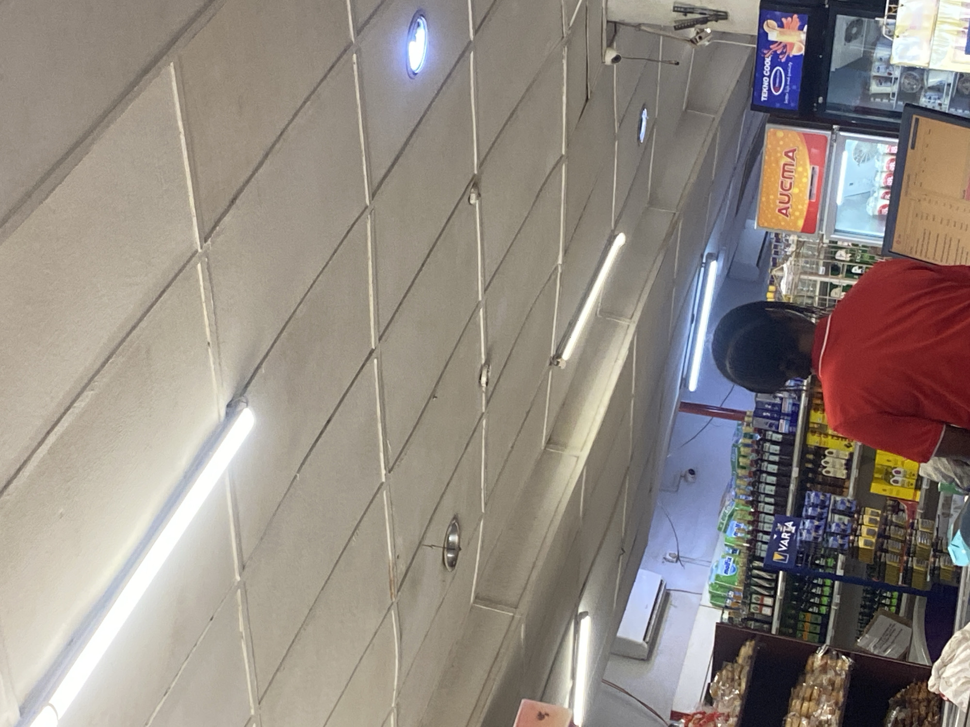 The media file shows the interior of a store in Ikorodu, Nigeria. The scene is illuminated by fluorescent strip lights and some ceiling lights. In the foreground, a person wearing a red shirt is partially visible, appearing to be a customer or employee. Behind them, shelves are stocked with various products, including what appears to be beverages and food items. Signs are visible above the shelves. One sign displays "TEKNO COOL" with a logo, and another shows "AUCMA" in orange and yellow lettering. Further back, a security camera is mounted on the ceiling. The overall impression is of a retail environment, likely a convenience store or supermarket. The lighting and the products on display suggest it is daytime or the store is well-lit. There are no strong indicators of the specific time of day beyond the artificial lighting. The general ambiance is that of a typical commercial setting.