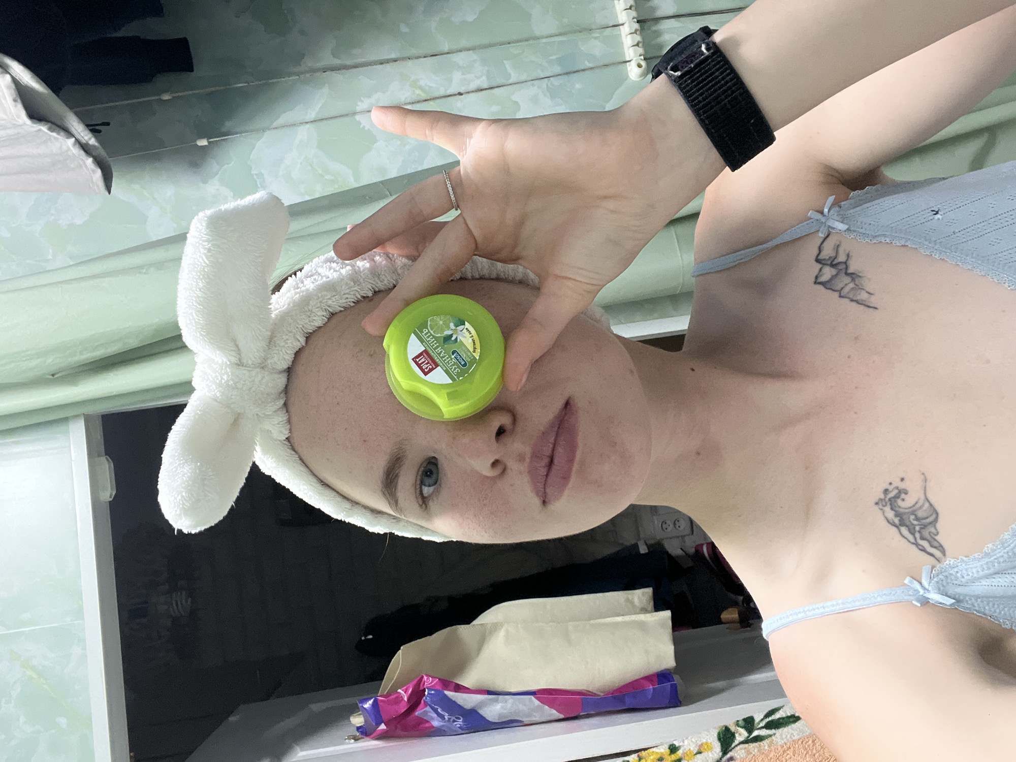 A young woman with fair skin, freckles, and light blue eyes is taking a selfie in a bathroom. She is wearing a white fluffy headband with a bow on top and a light blue camisole. She is holding a small, round, green container of dental floss in front of her right eye. The container has text and an image of lime and a flower. Her left hand is raised, and she is wearing a thin silver ring on her left ring finger and a black watch on her right wrist. A tattoo of a wave is visible on her left chest, and a tattoo of mountains is visible on her right chest. Behind her, a white cabinet door is open, revealing clothes and bags. A green curtain hangs in the background. The lighting suggests an indoor environment, possibly a residential bathroom. The city of Chaikovsky, Russia, is not discernible from the image.